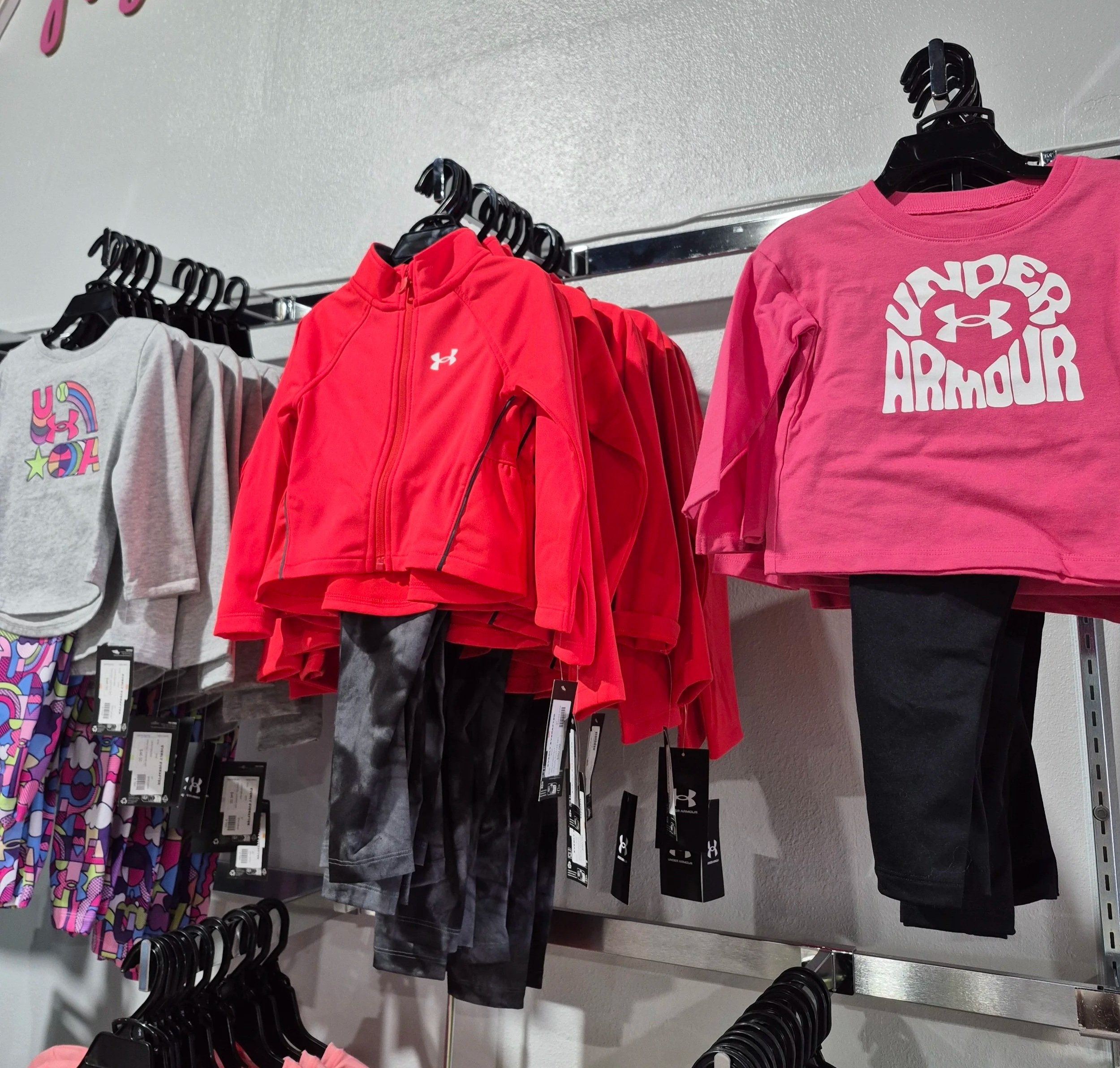 Kids' athletic clothing displayed on store racks, including a pink Under Armour crop top, a red Under Armour jacket, and gray and patterned leggings.