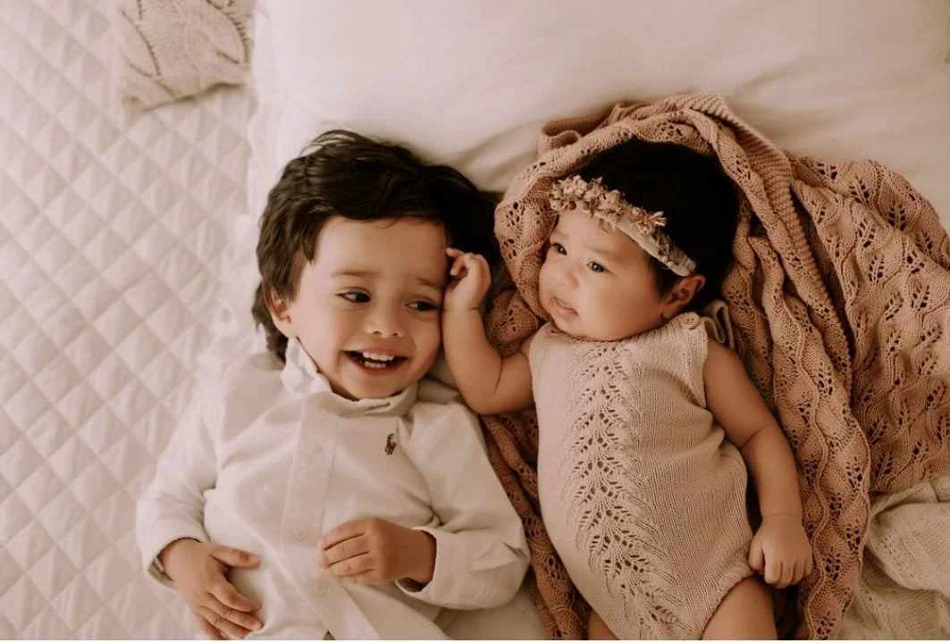 A young child smiling next to a baby on a bed, with the baby wrapped in a knitted blanket.
Everly Everafter Kids | Children's Clothing Store