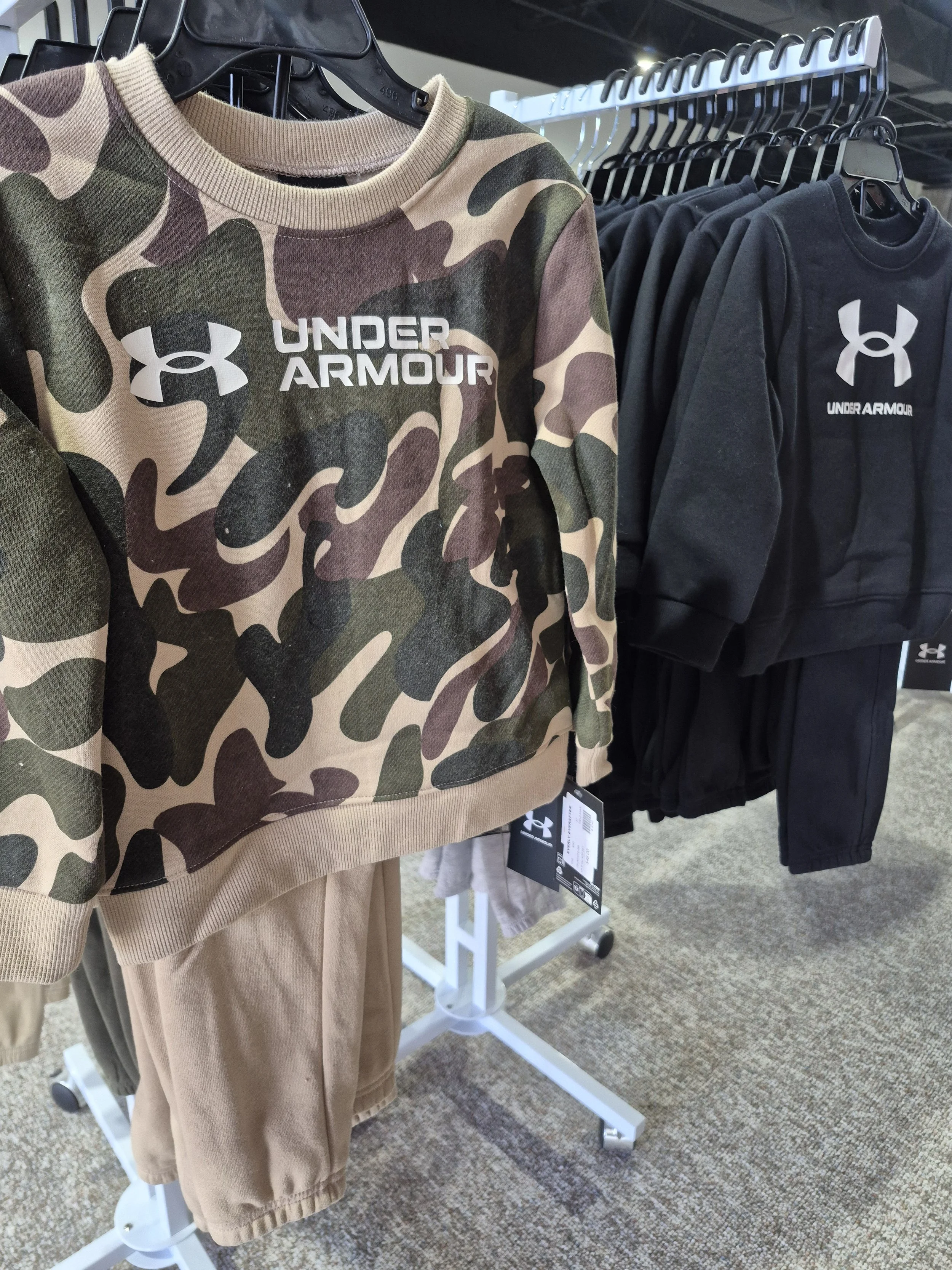 A camouflage Under Armour sweatshirt with beige, green, and brown colors hanging on a clothing rack, alongside black sweatshirts with the Under Armour logo. Beige pants are partially visible below the sweatshirt.