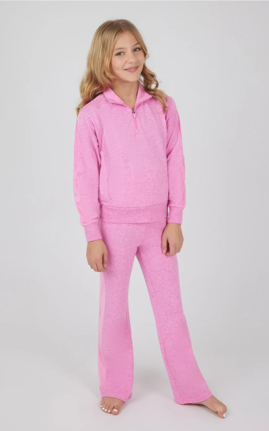 A young girl with blonde hair wearing a pink speckled tracksuit, standing against a plain light gray background.