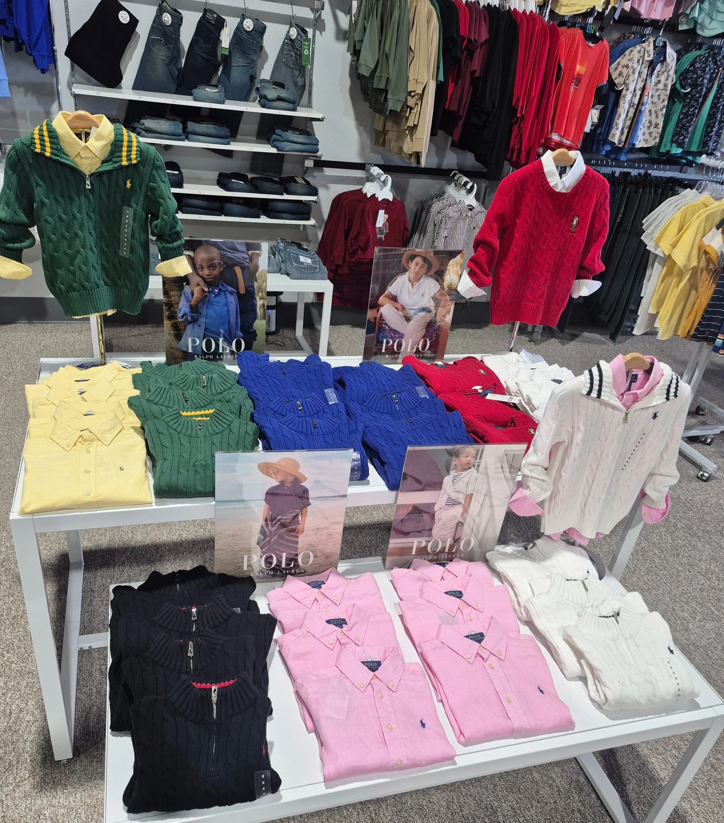 Display of colorful sweaters and shirts in a clothing store, with promotional posters of children modeling the apparel.