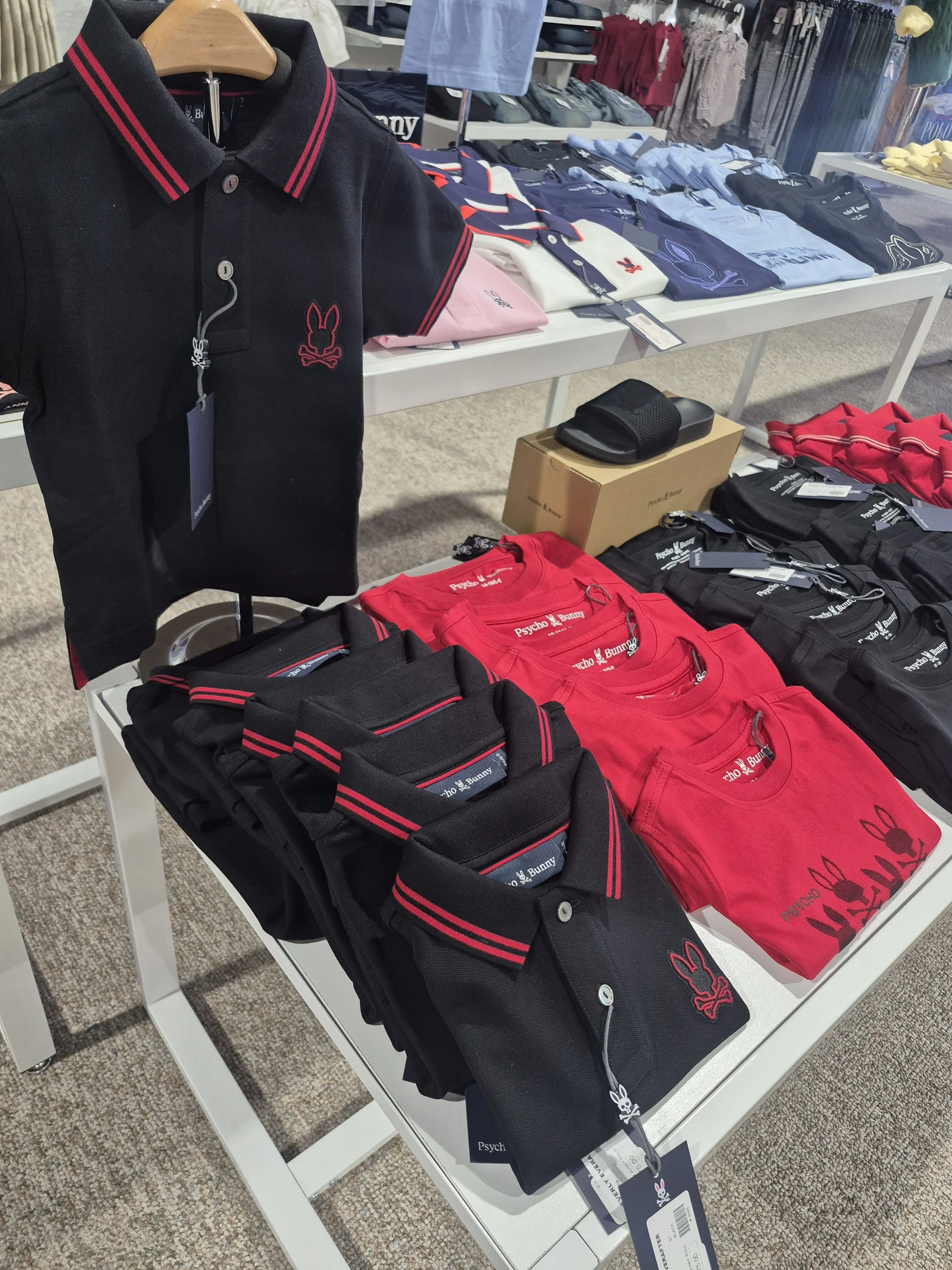 Table displaying folded and hanging clothing items, including black, red, and light blue shirts with a bunny logo, in a retail store.