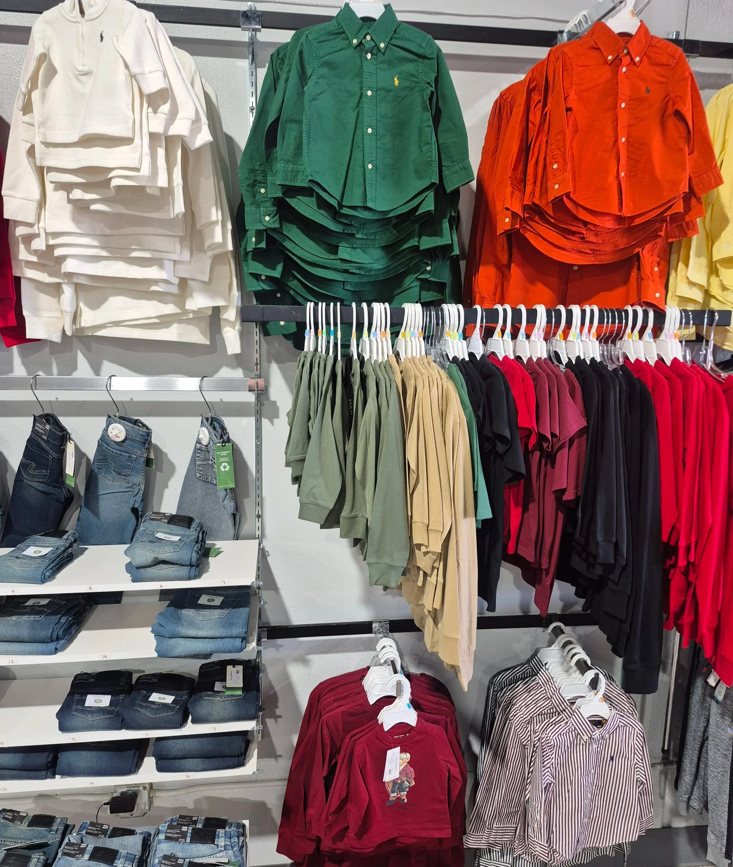 Display of various clothing items in a retail store, including white hoodies on the left, green and orange button-up shirts hanging above, rows of assorted hoodies in green, beige, black, red, and other colors, jeans on the lower left shelf, and red and striped shirts hanging below.