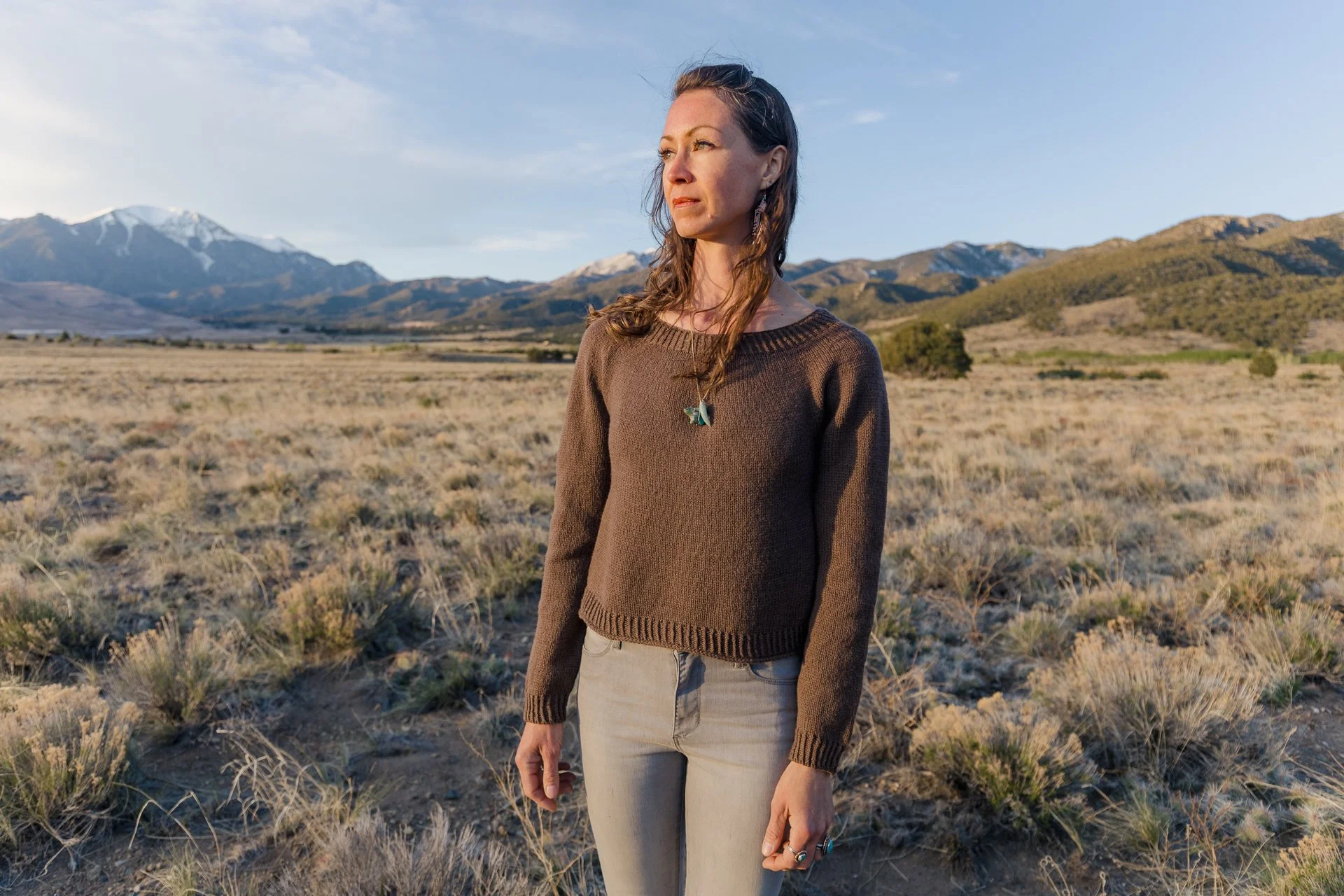 A woman standing outdoors in a dry, grassy field with mountains in the background, wearing a brown sweater and beige pants, looking into the distance during sunset.