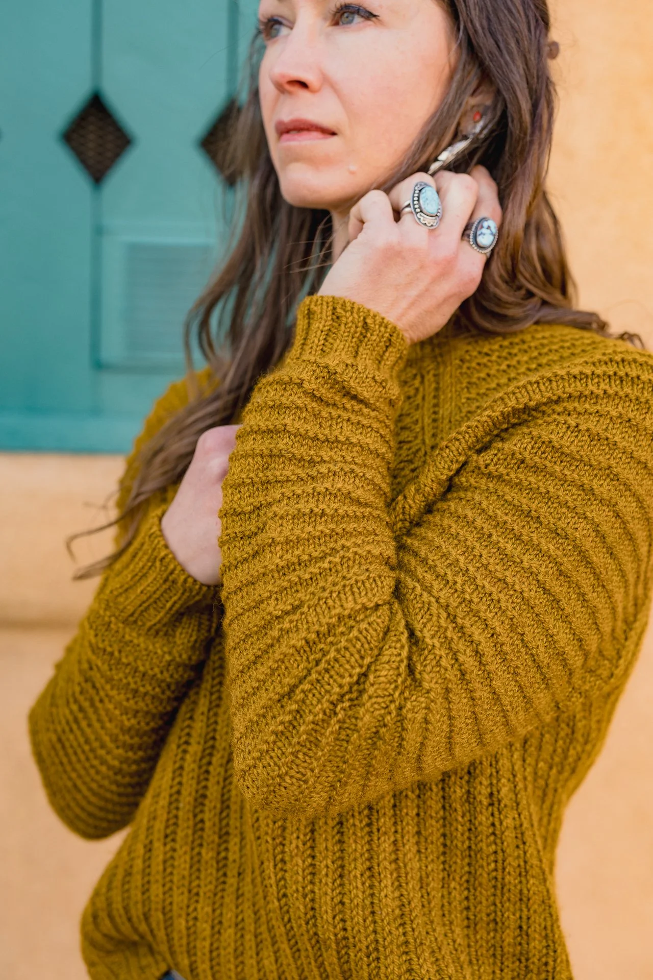 A woman with brown hair wearing a mustard yellow hand knit brioche and garter stitch sweater with turquoise rings standing in front of a teal door.