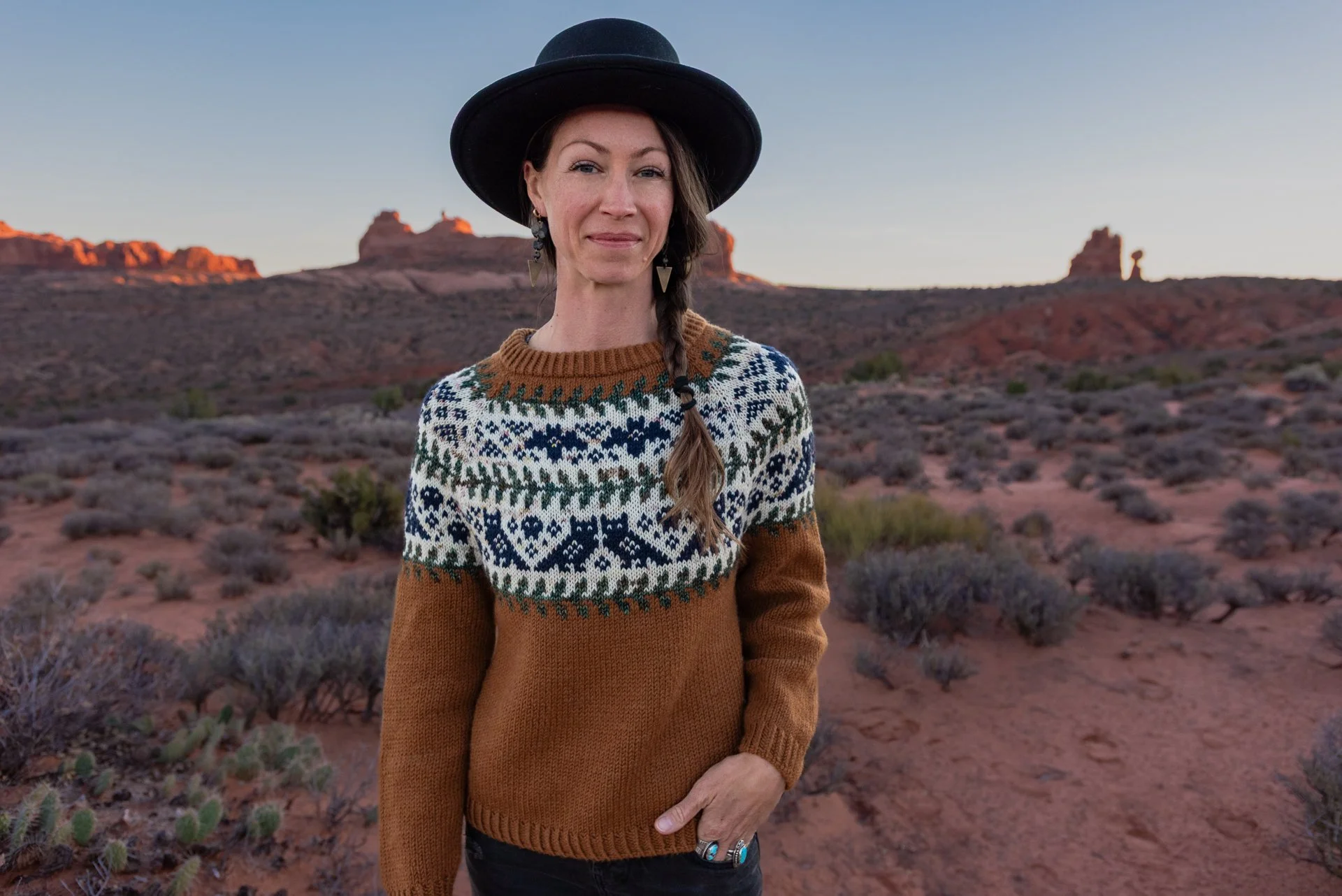 A woman with long braided hair and earrings, wearing a black hat and a colorful knit sweater, standing in a desert landscape with red rock formations at sunset.