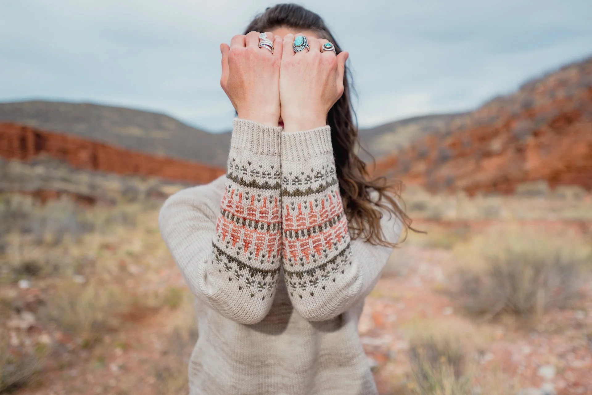 Woman standing outdoors in a desert landscape with mountains, covering her face with her hands, wearing a cream-colored sweater with colorful knit patterned sleeves and rings on her fingers.