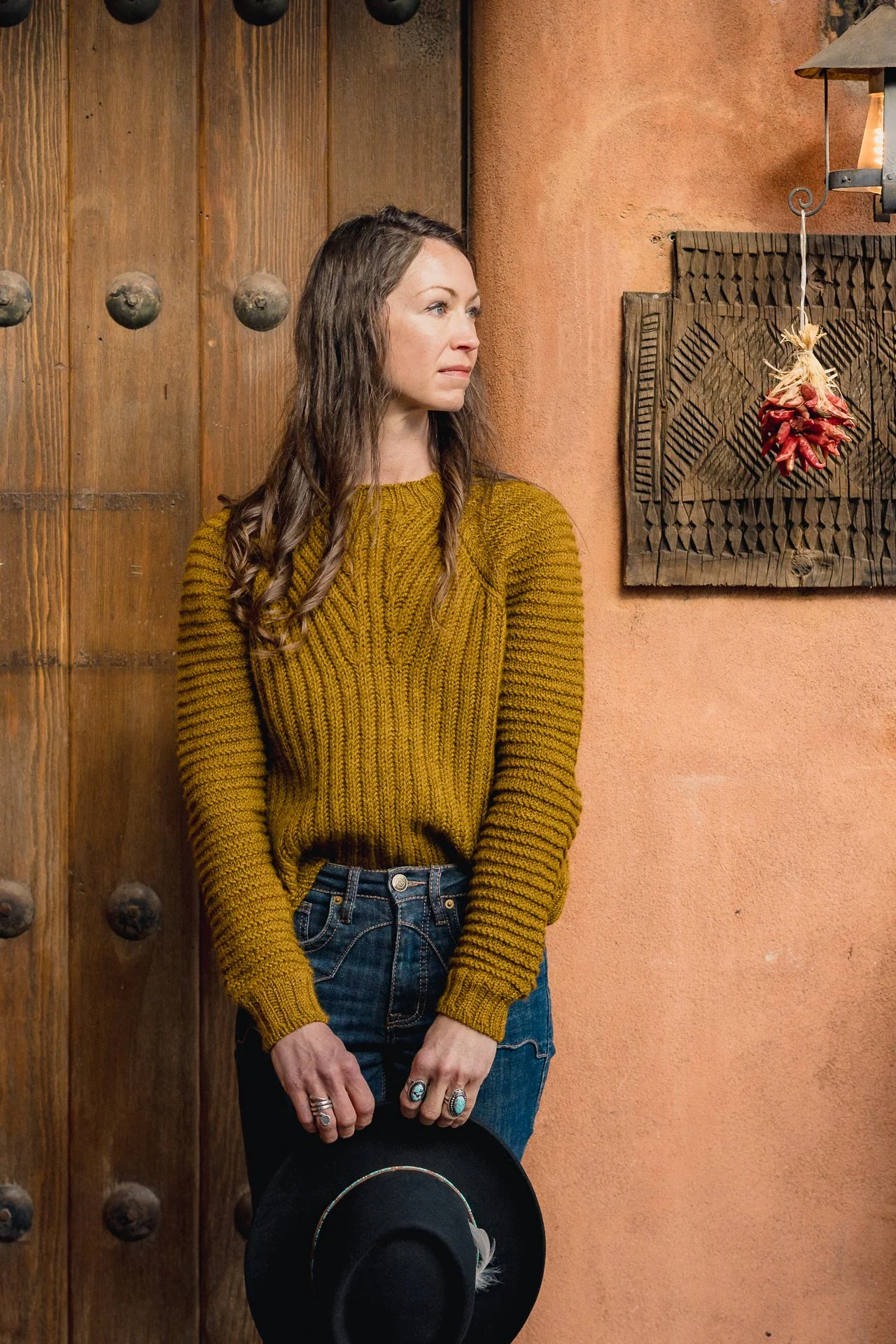 A woman with long, wavy hair wearing a yellow hand knitted brioche sweater, blue jeans, and turquoise rings. She is holding a black hat in her right hand and standing against a rustic background of wooden and adobe walls.