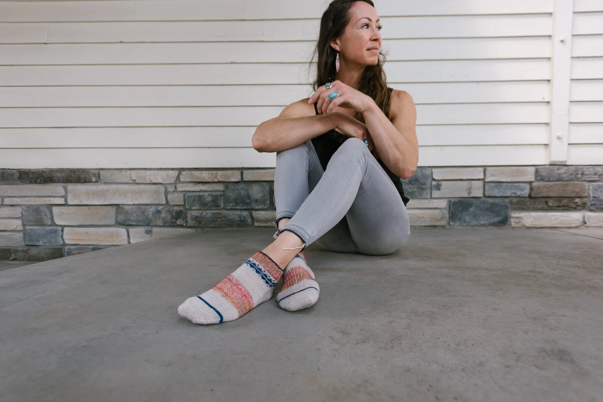 A woman sitting on the ground outdoors with her back against a wall, looking to the side, wearing a black tank top, gray jeans, colorful socks, and earrings.