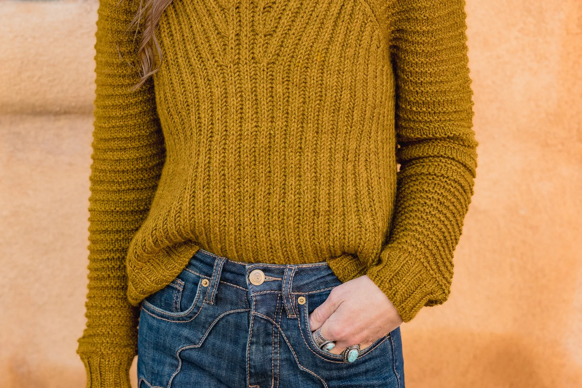 Person wearing a mustard yellow chunky knit brioche sweater with long sleeves and a round neck, paired with blue denim jeans that have prominent stitching and a button closure, with their left hand resting in the pocket.
