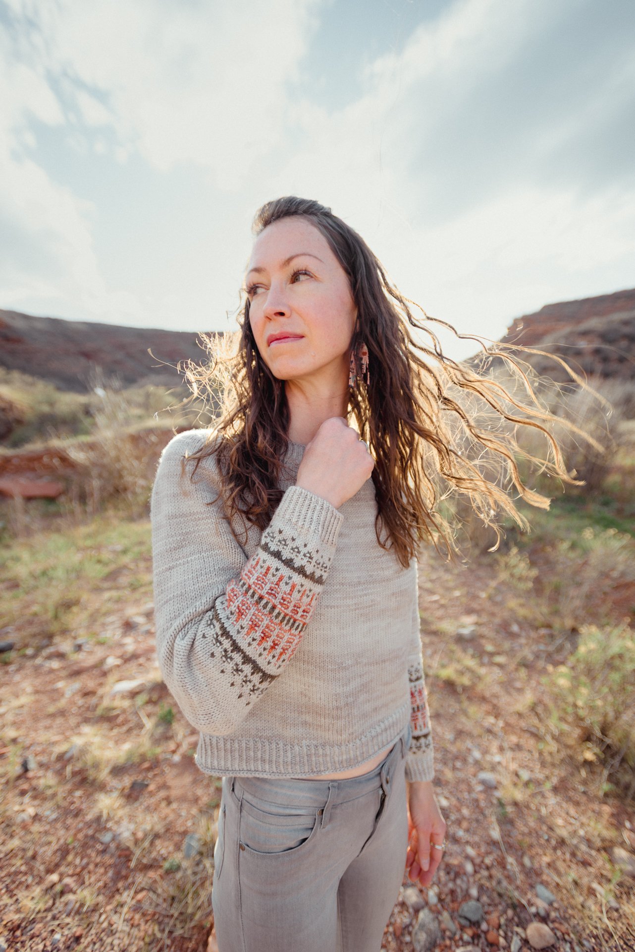 A woman with long curly hair standing outdoors in a natural landscape with hills and sparse vegetation, wearing a beige sweater with patterned sleeves and gray pants, looking off into the distance with windblown hair.
