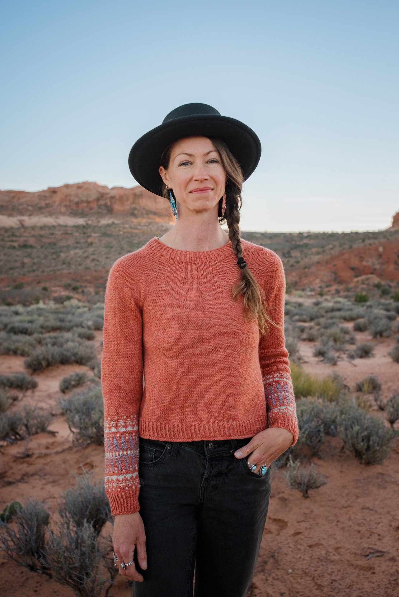 A woman with a braid, wearing a black hat, orange handknit sweater featuring stranded colorwork on the lower sleeves, and black pants, standing in a desert landscape with mountains and sparse shrubs.