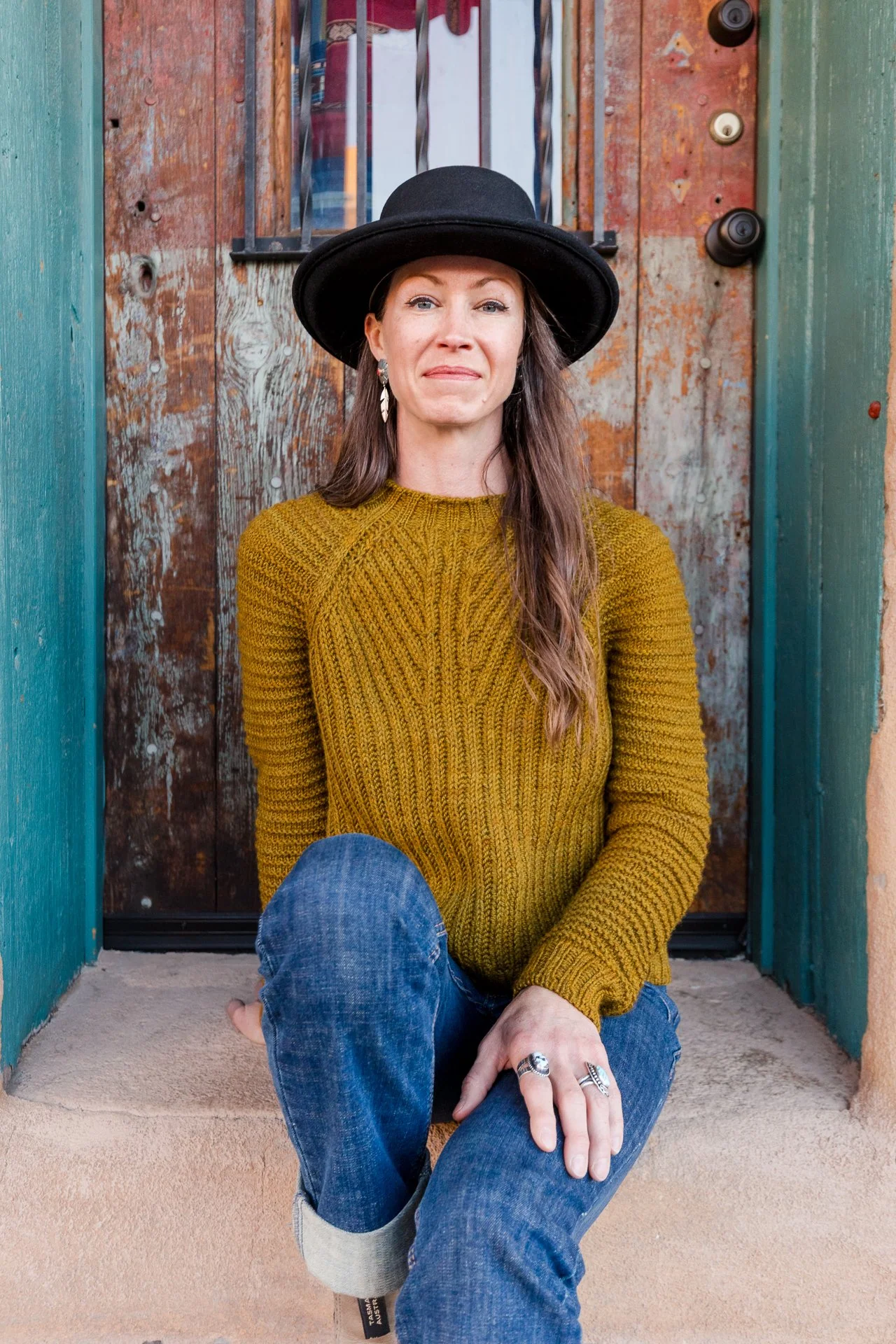 A woman with long brown hair, wearing a black hat, yellow hand knit brioche sweater, and blue jeans, sitting on a porch with a weathered wooden door behind her.