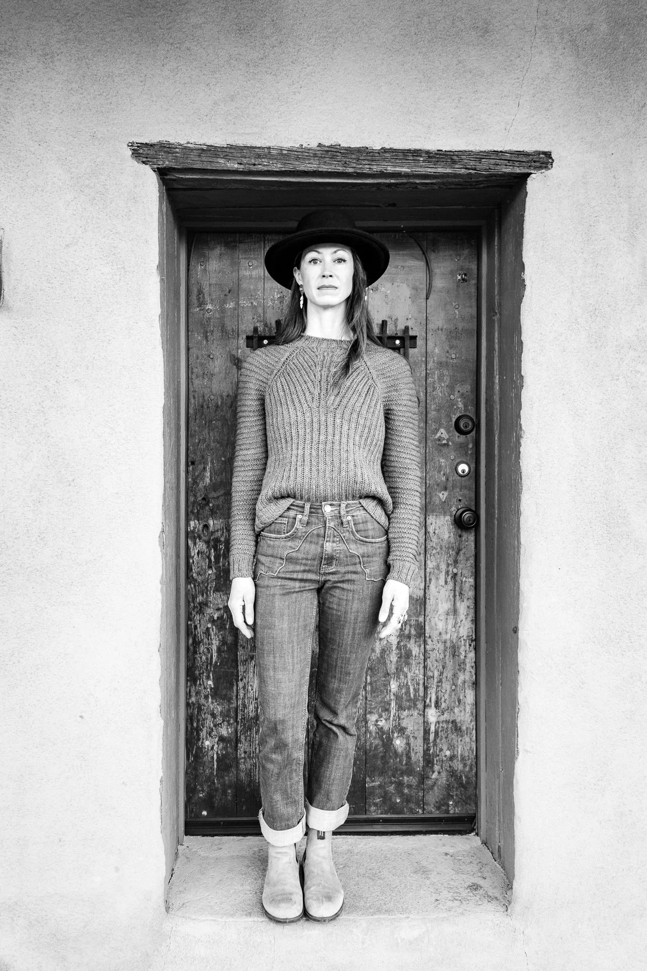 A woman standing in a doorway outside, wearing a wide-brimmed hat, hand knitted textured brioche sweater, jeans, and boots, in black and white photo.