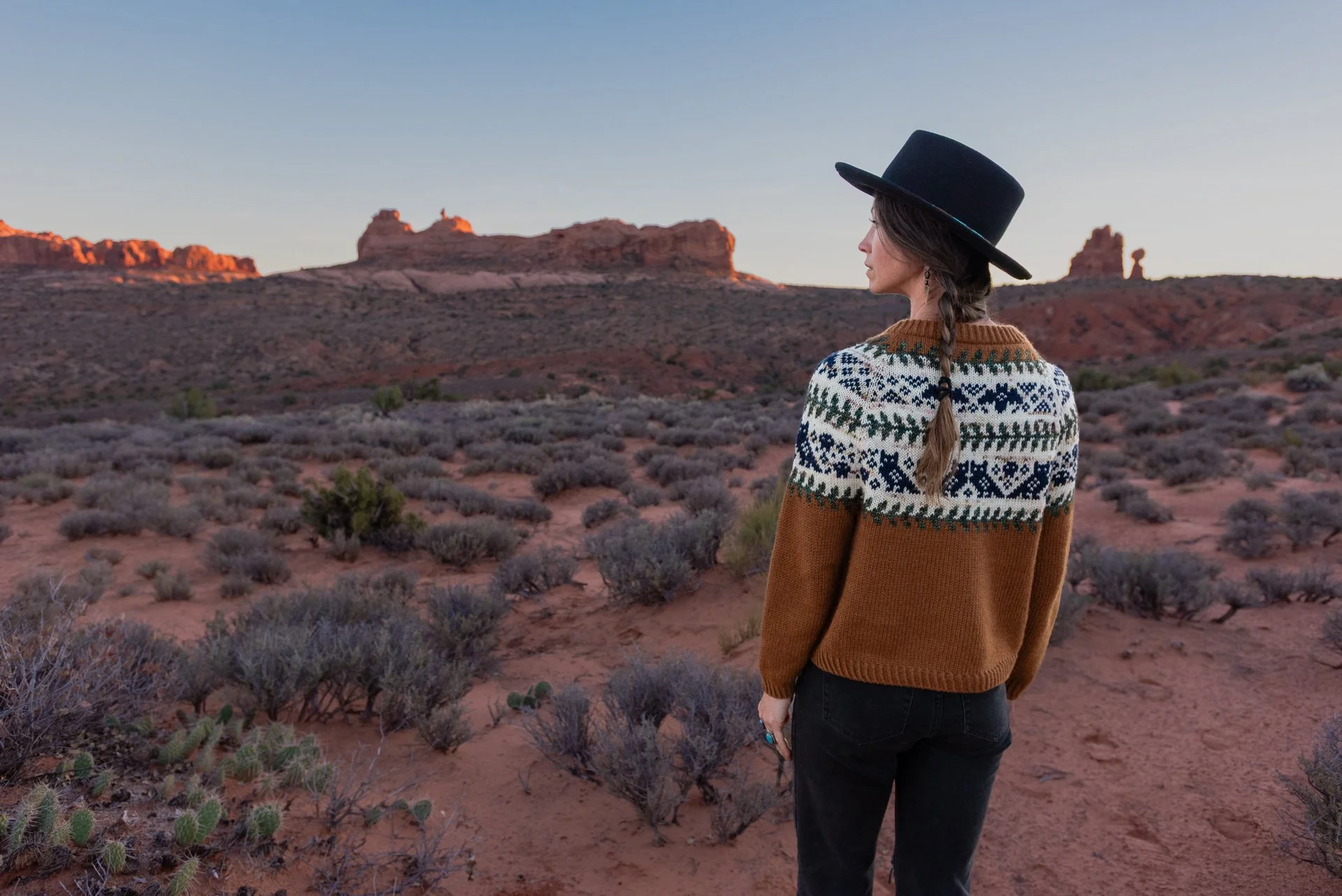 Woman in a patterned sweater and black hat standing in desert landscape with red rock formations and sparse vegetation at dusk.
