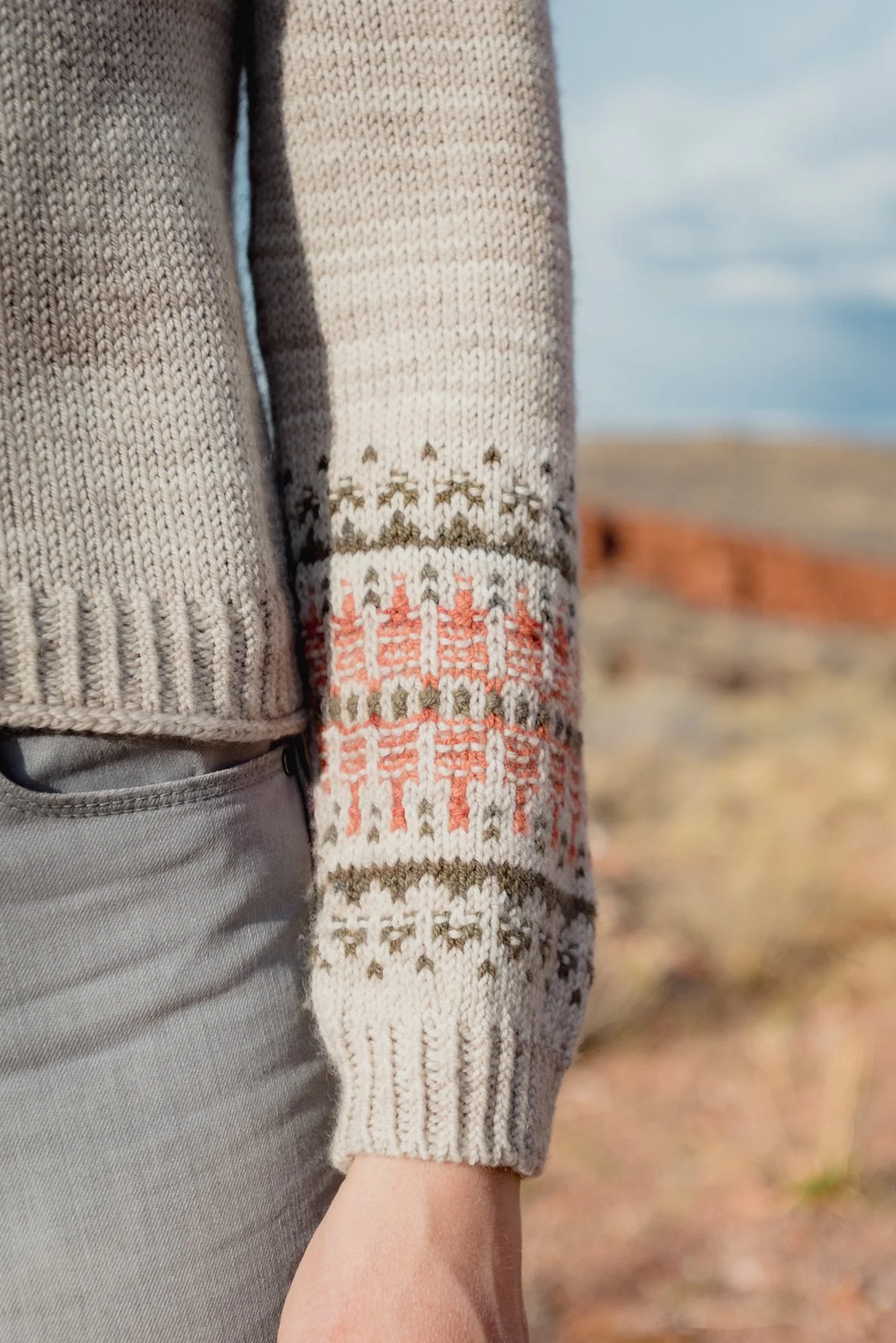 Close-up of a person wearing a beige patterned sweater with embroidery, beige pants, and a glimpse of a hand near the pocket. The background shows an outdoor landscape with land and water under a partly cloudy sky.