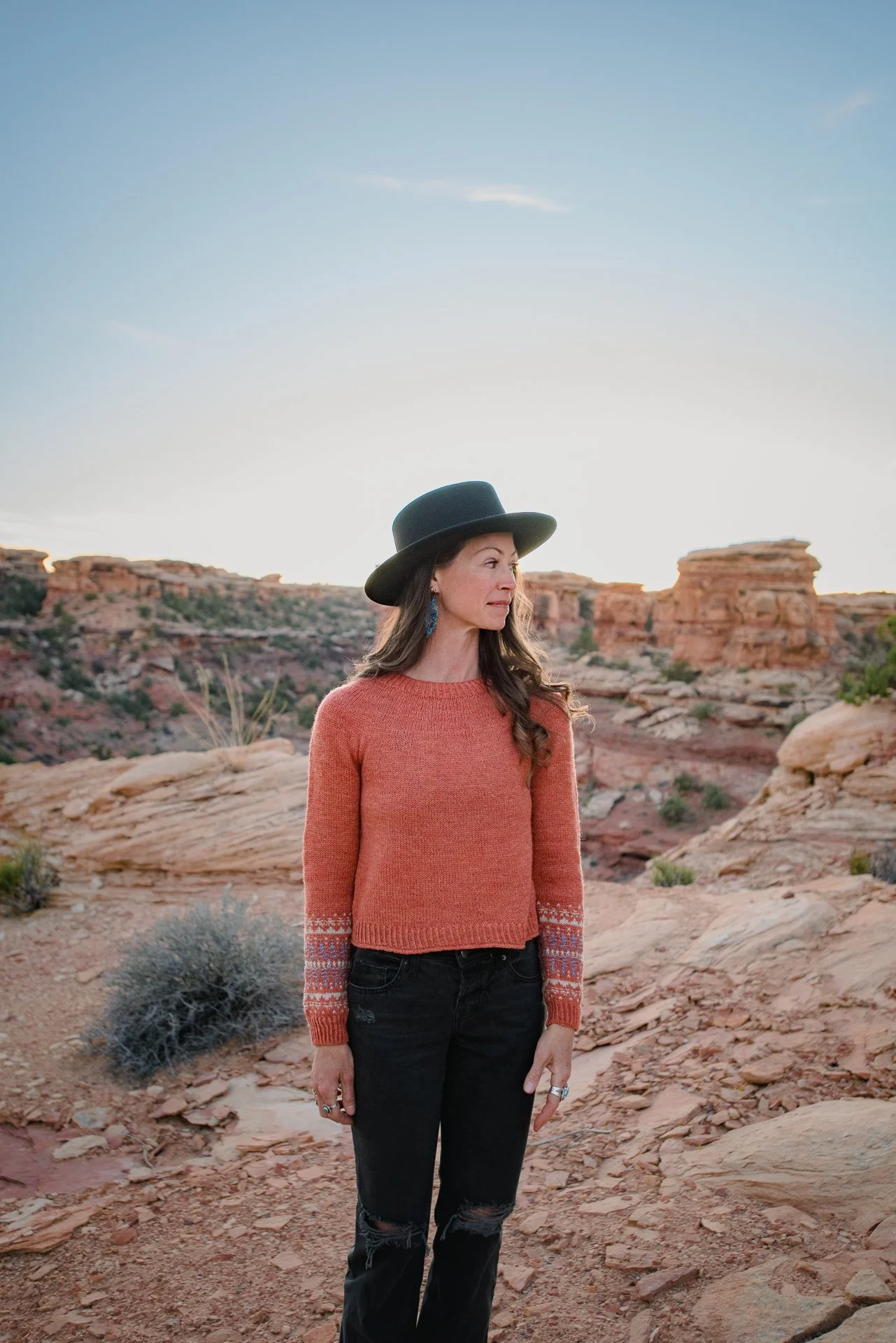 A woman in a coral sweater and black torn jeans standing outdoors in a desert landscape with rock formations at sunset, wearing a black hat and jewelry.