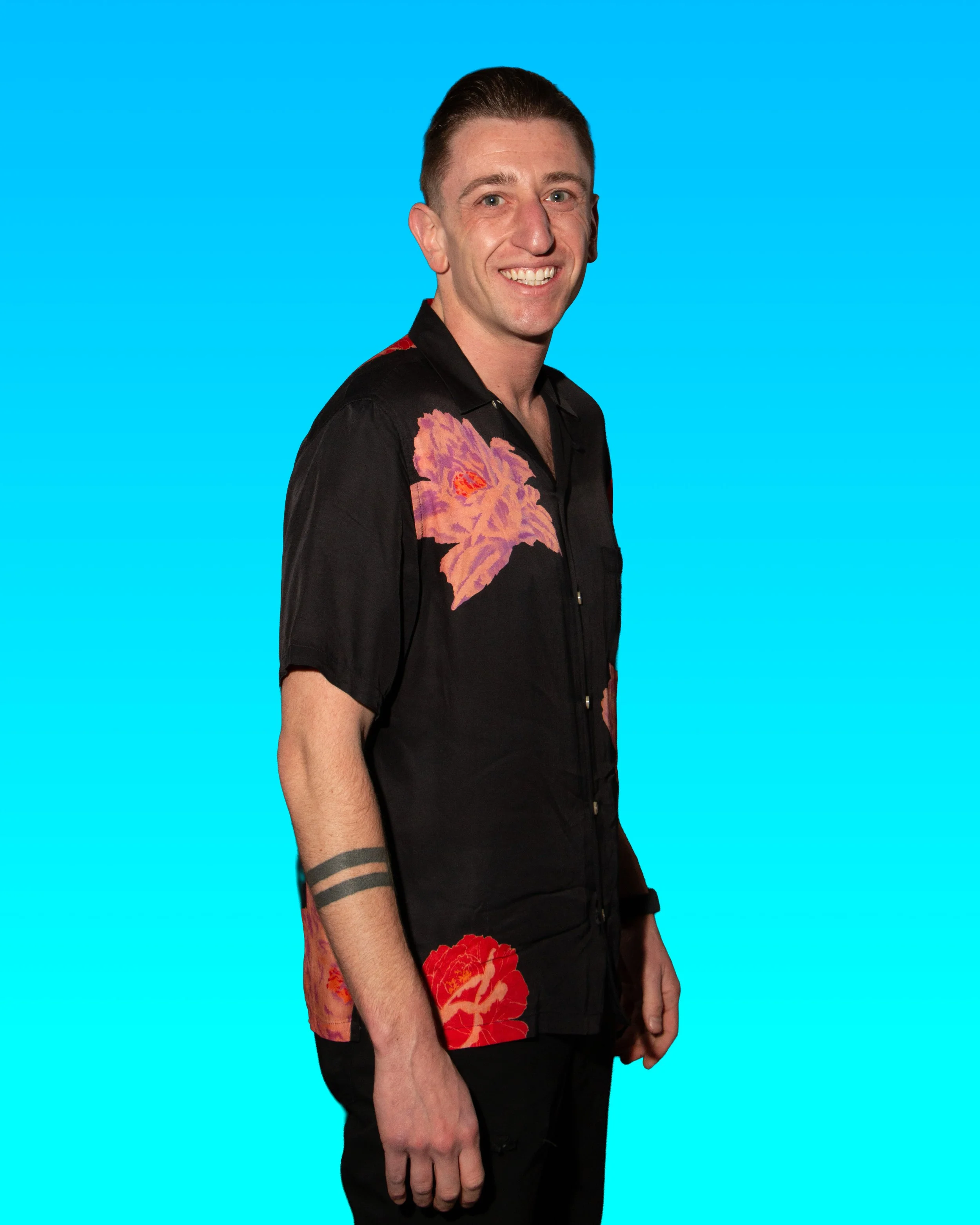 Young man smiling, wearing a black shirt with red and pink floral design, standing against a bright blue background.