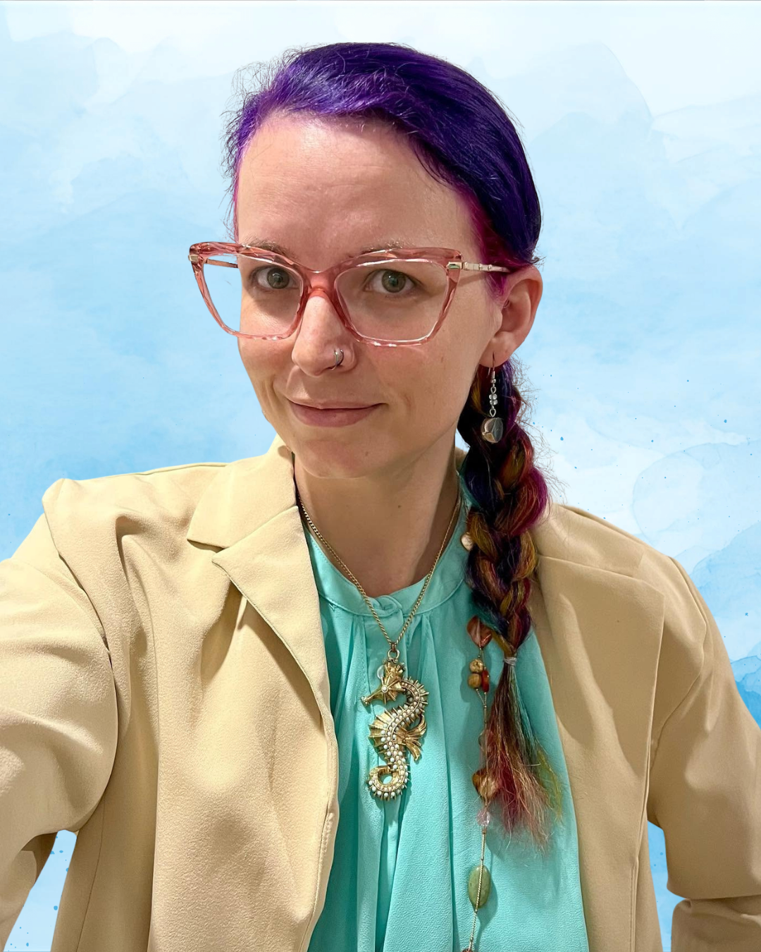 A woman with purple and pink braided hair wearing pink glasses, a beige blazer, and a turquoise blouse, accessorized with a seahorse necklace and colorful earrings, standing against a light blue background.