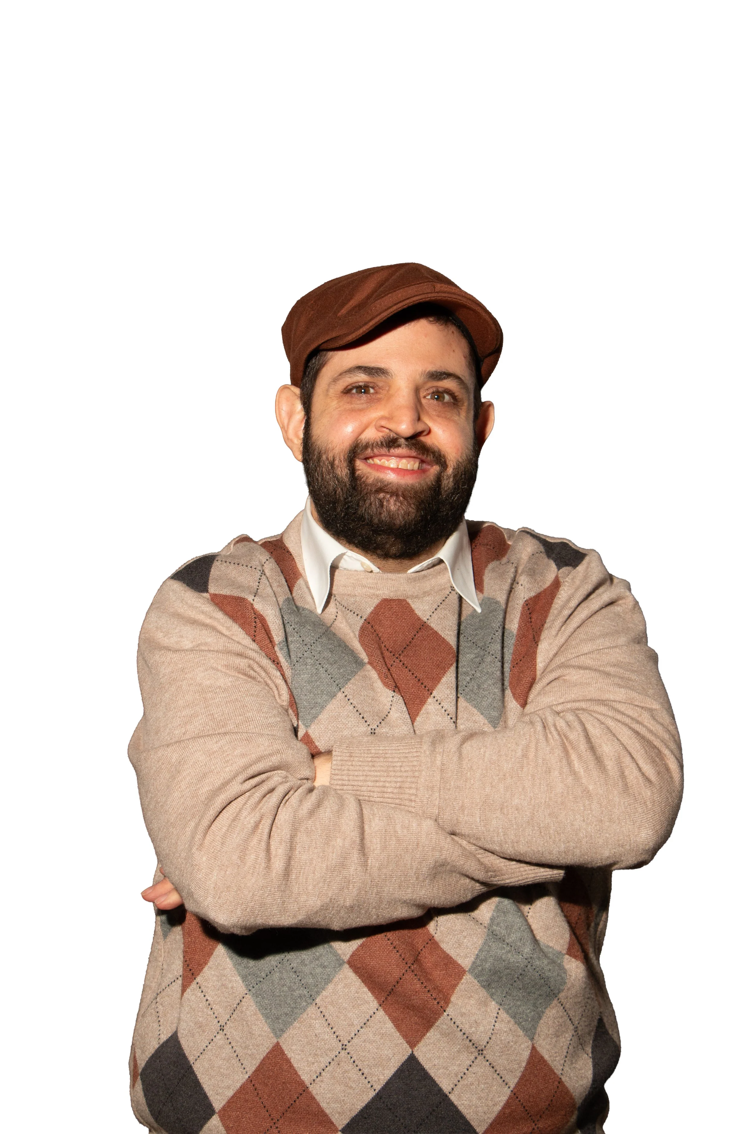 A man with a beard and short dark hair, wearing a brown cap and a beige argyle sweater, smiling with crossed arms against a white background.