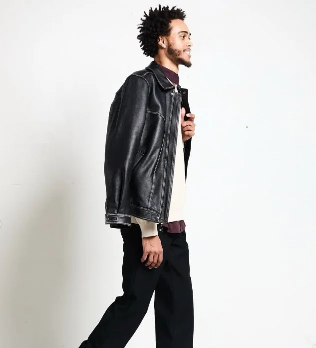 A man with curly hair and a beard walking to the right, wearing a black leather jacket, black pants, and a white shirt against a plain white background.