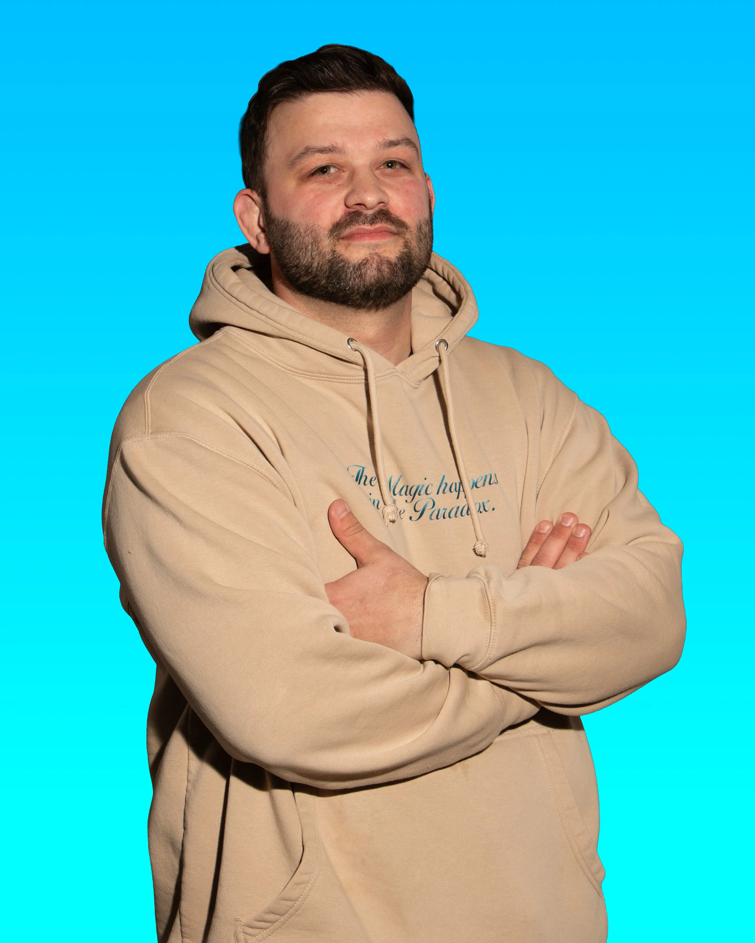 A man with a beard and short dark hair, wearing a beige hoodie with blue text, stands with his arms crossed against a bright blue background.