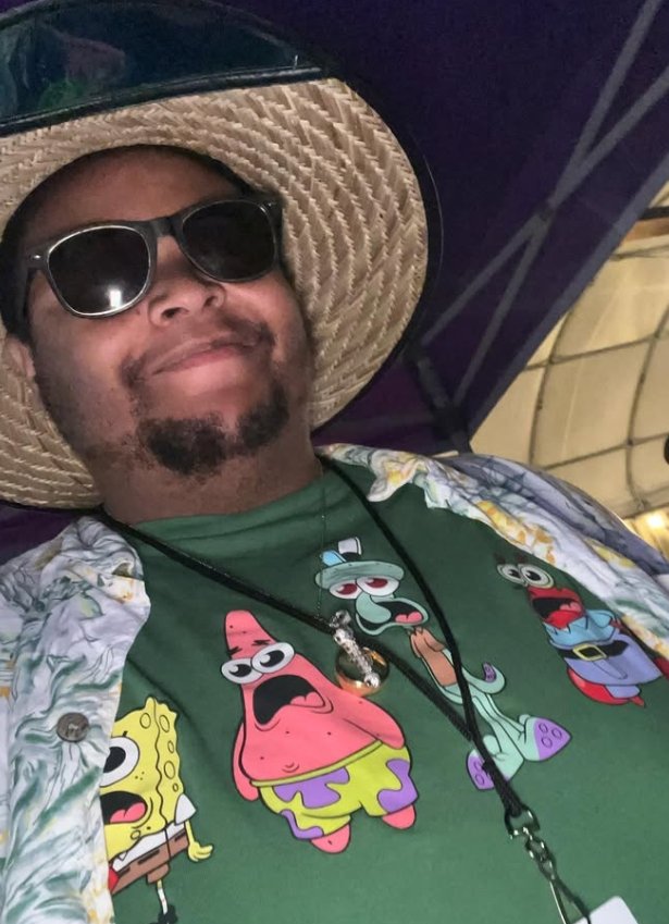 A man wearing a wide-brimmed straw hat, sunglasses, and a festive shirt with cartoon characters from SpongeBob SquarePants, including SpongeBob, Patrick, Squidward, and Mr. Krabs, smiling at the camera.