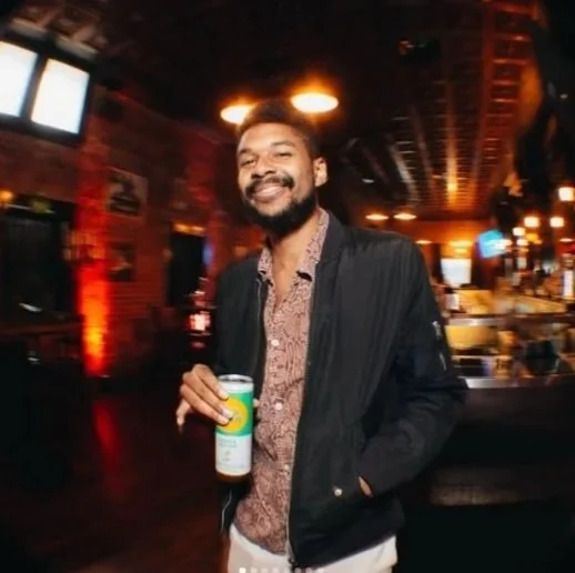 A smiling man holding a can in a dimly lit bar or pub with warm lighting and wooden decor.