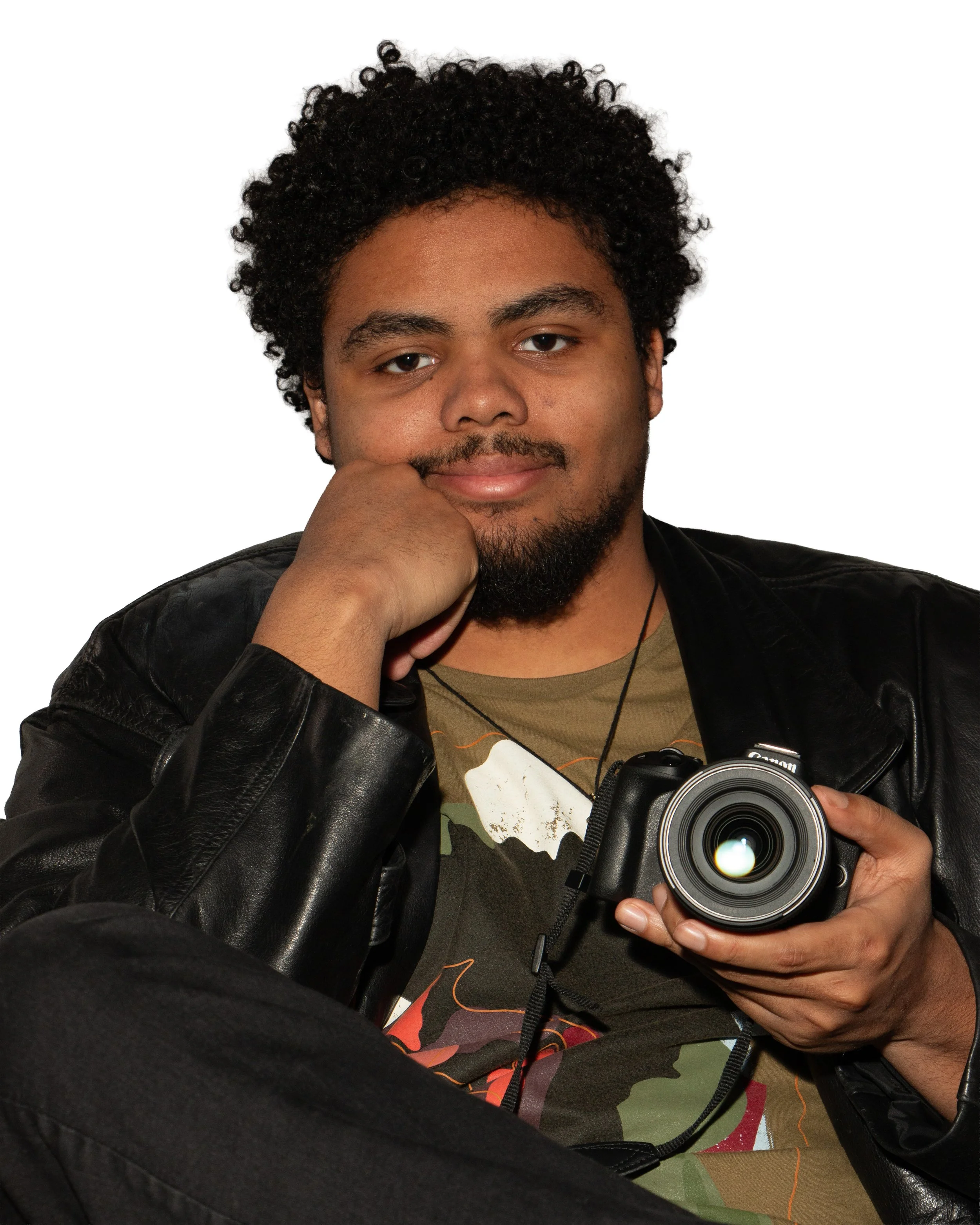 A young man with curly black hair and a beard, wearing a black leather jacket and a graphic t-shirt, is holding a camera and taking a selfie. He is smiling slightly and looking into the camera, with his right hand resting on his chin in a resting pose, against a plain white background.