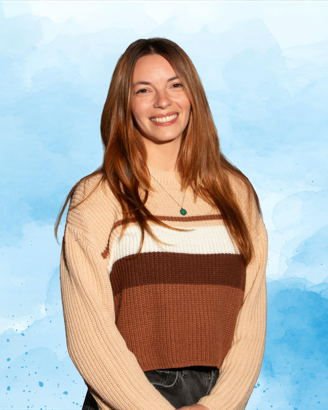 A young woman with long red hair, smiling, wearing a beige, brown, and white striped sweater and a pendant necklace, standing against a blue textured background.