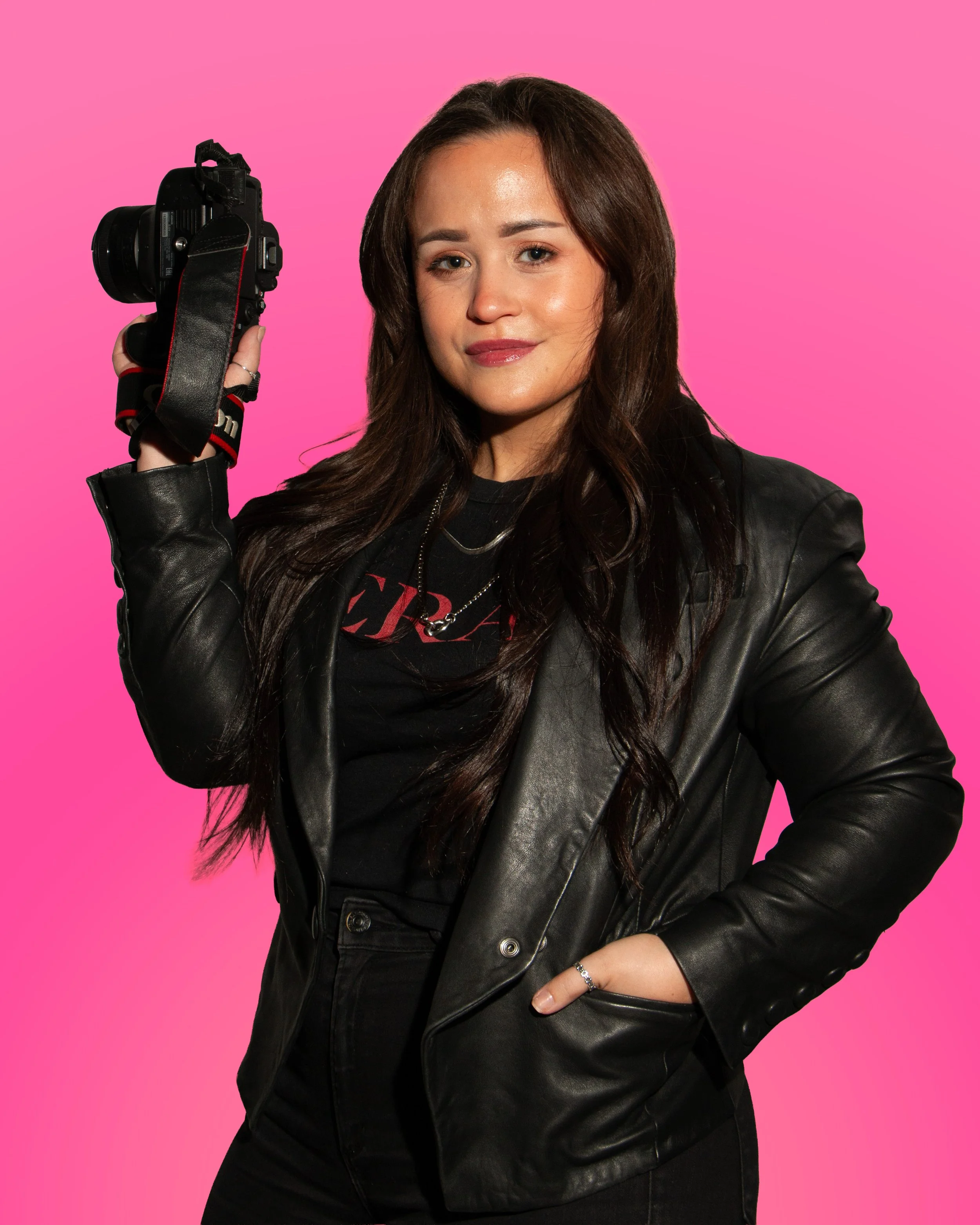 A woman with long brown hair wearing a black leather jacket and black shirt, holding a camera, against a pink background.