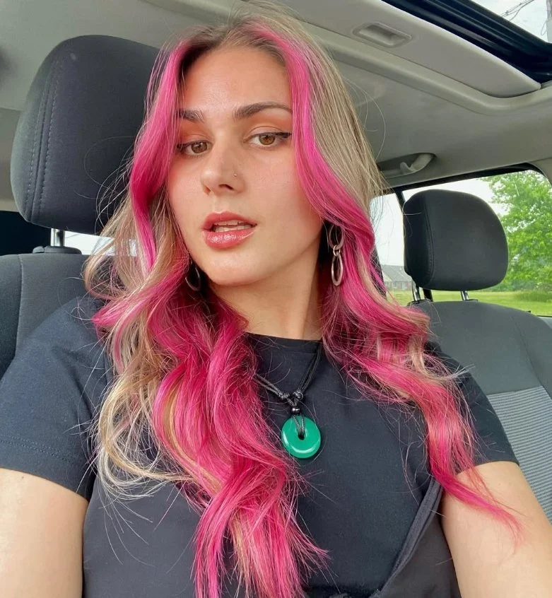 A young woman with long, wavy hair dyed pink and blonde, sitting in a car. She has a nose piercing, hoop earrings, and is wearing a black t-shirt with a green pendant necklace. Outside the car window, green trees are visible.