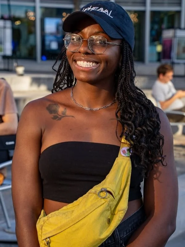 A smiling woman with glasses, long braided hair, and a tattoo of a flower on her left shoulder, wearing a black tube top, a yellow bag across her body, a necklace, and a blue cap, outdoors with people and a building in the background.