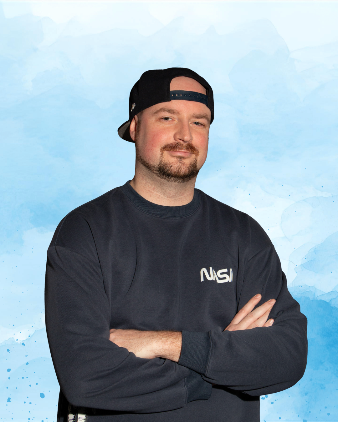 A man with a goatee and mustache wearing a black NASA sweatshirt and a backwards black baseball cap against a blue watercolor background.