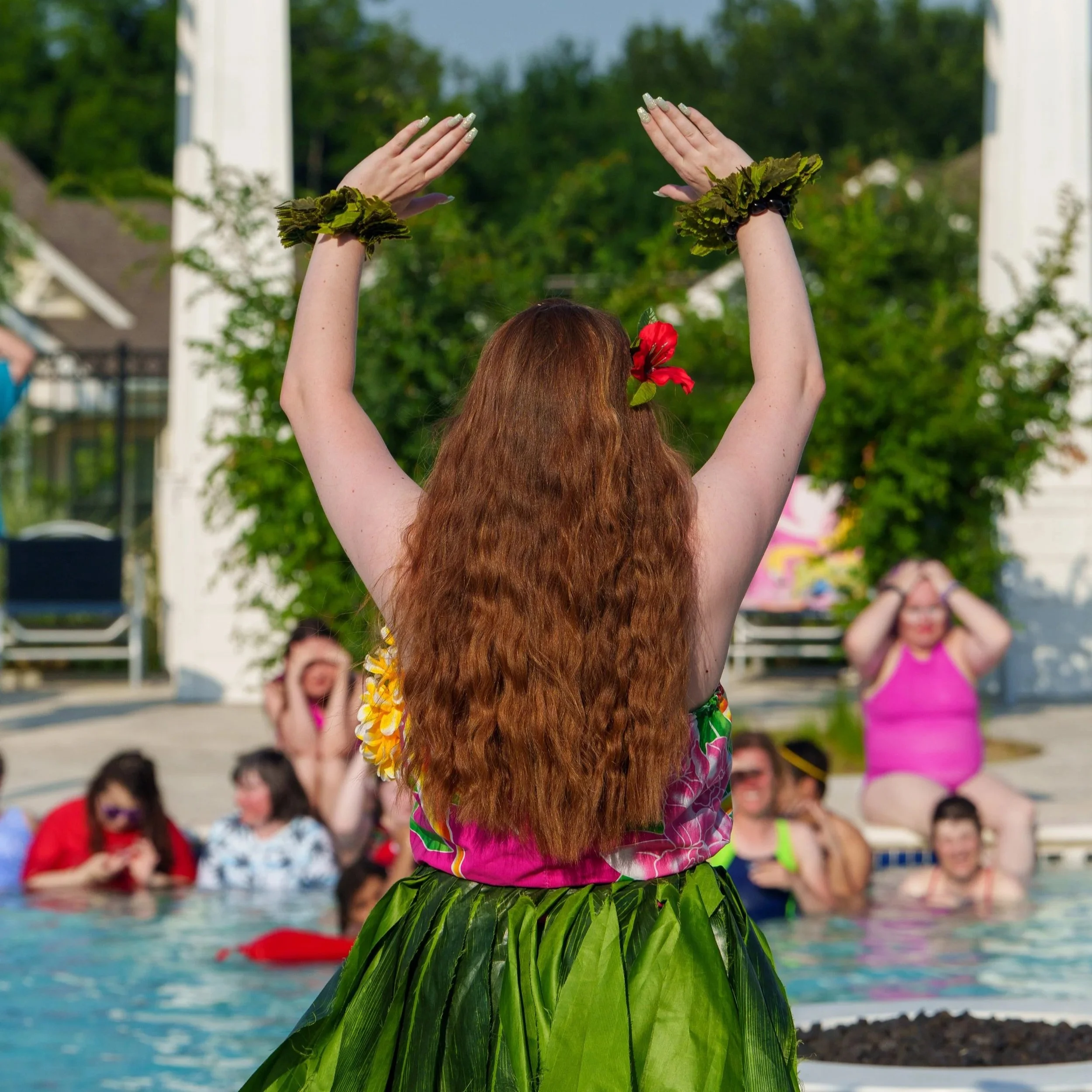 Person with long red hair wearing a hula skirt and floral accessories performing a dance by a swimming pool, with an audience in the background.