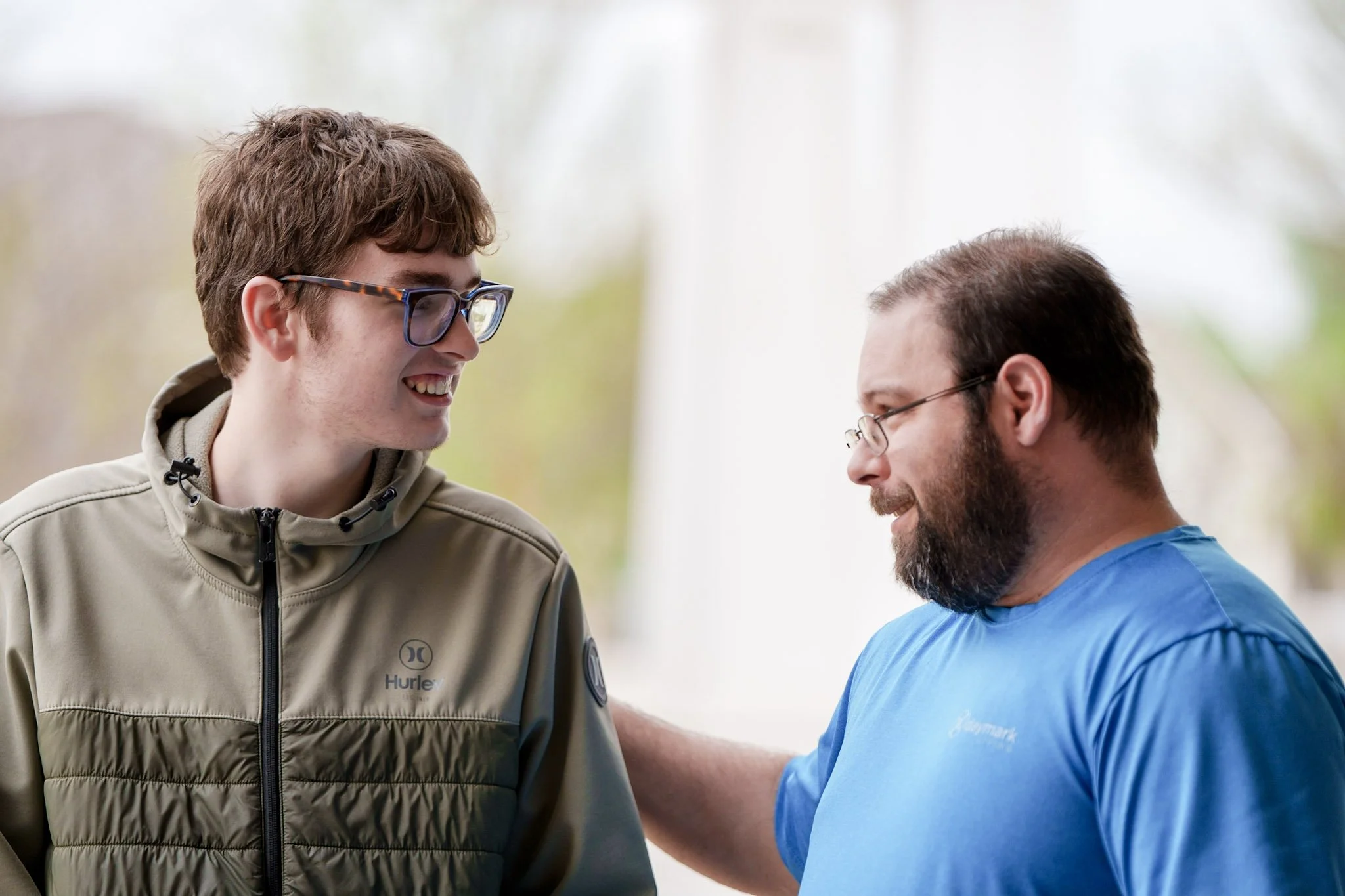 Two men smiling and talking outdoors, one with glasses and wearing a beige jacket, the other with glasses and a blue shirt, with trees in the background.