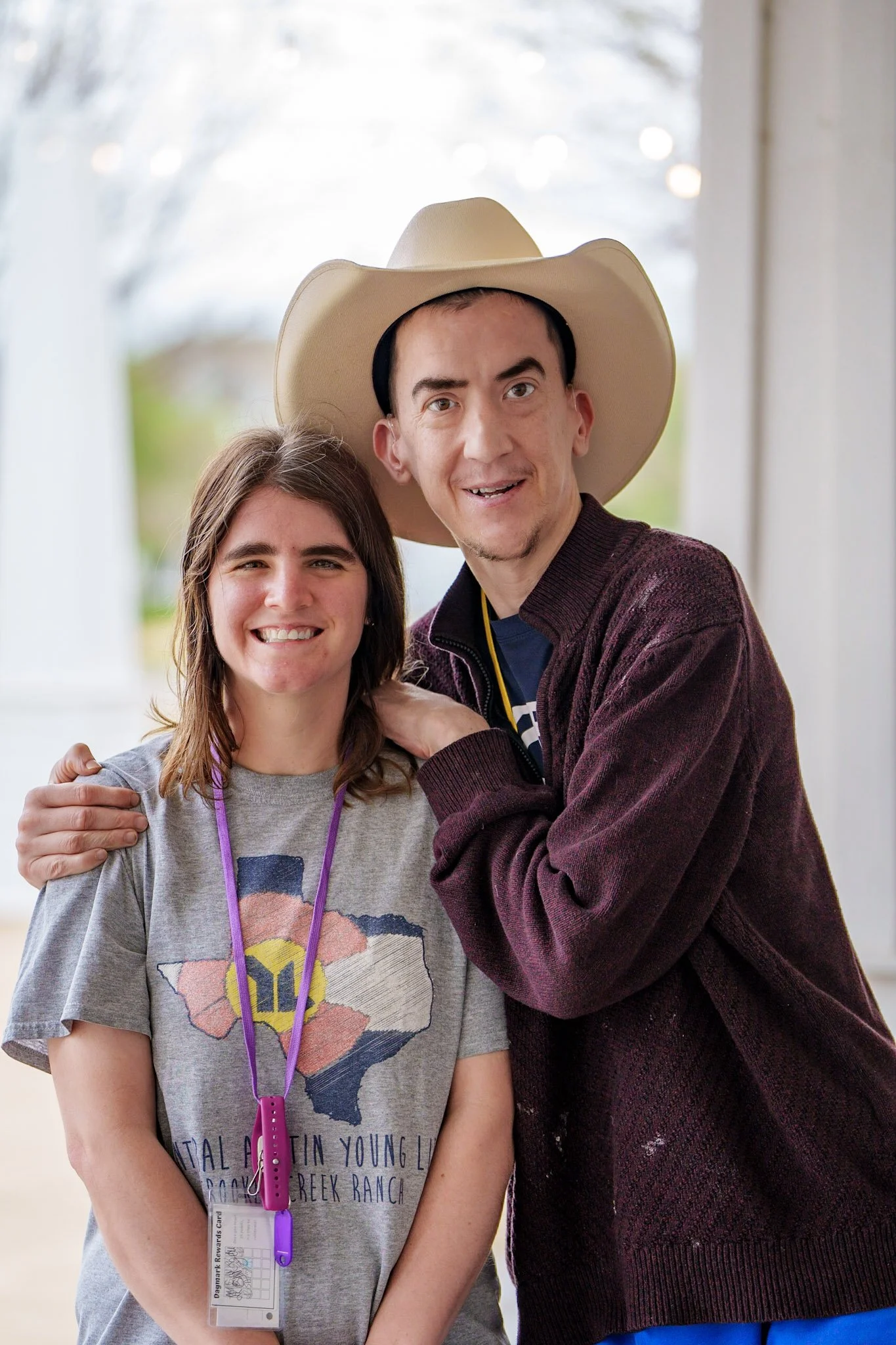 A young woman and a man, both smiling, standing close together outside. The man is wearing a large cowboy hat and a burgundy cardigan, and the woman is wearing a gray T-shirt with a Texas-themed design.