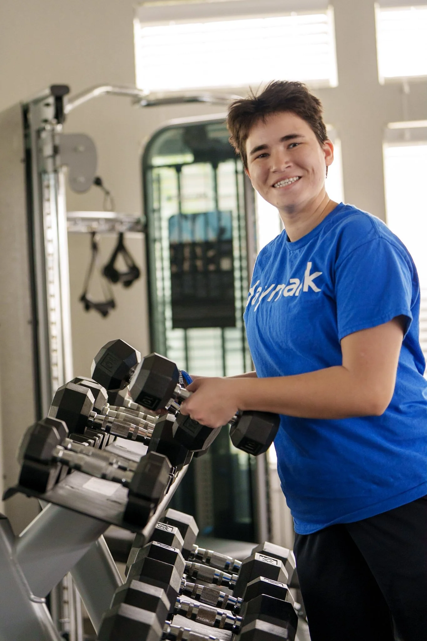 A young person smiling while lifting a dumbbell in a gym, wearing a blue shirt with white text.
