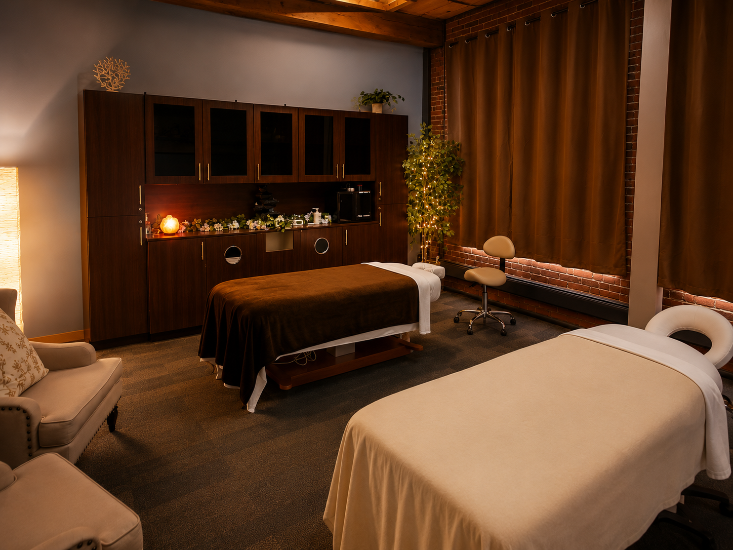 A cozy massage therapy room with two massage tables, one with a brown cover and the other with a white cover, in front of a wooden cabinet with plants and decorative items. There is warm lighting, beige curtains, and a brick wall background.