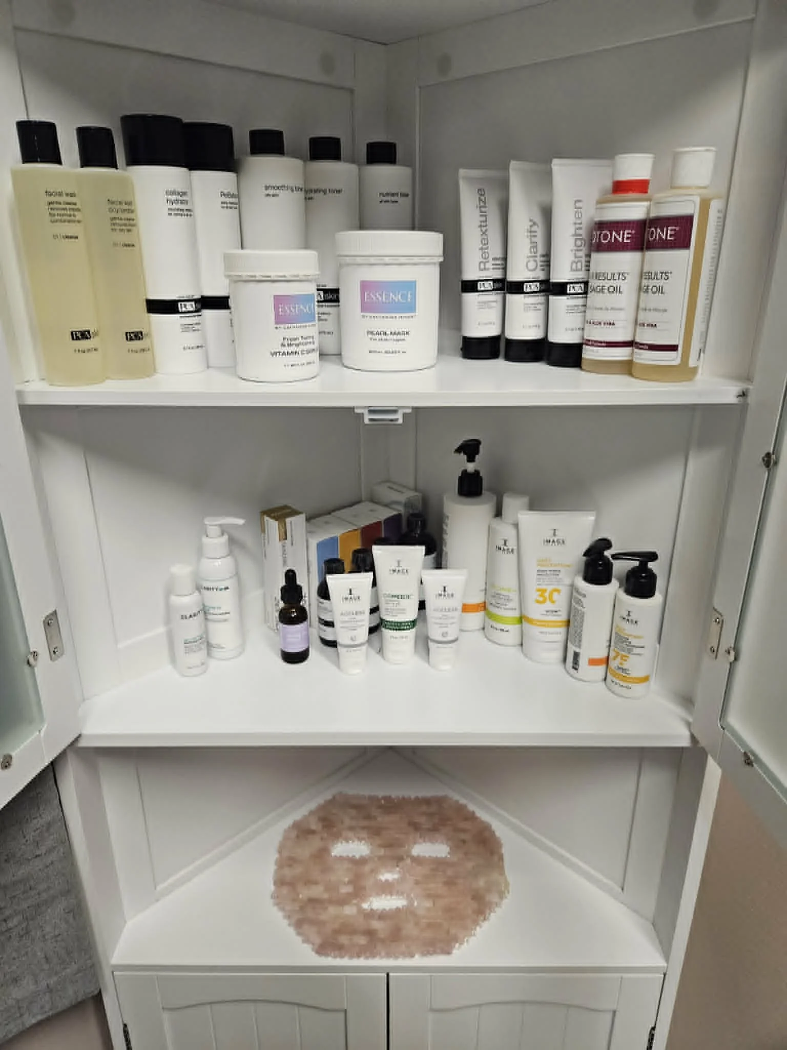 A white cabinet with three shelves holding skincare and hygiene products, including bottles of lotion and cream, with a heart-shaped pink plush rug on the floor in front.