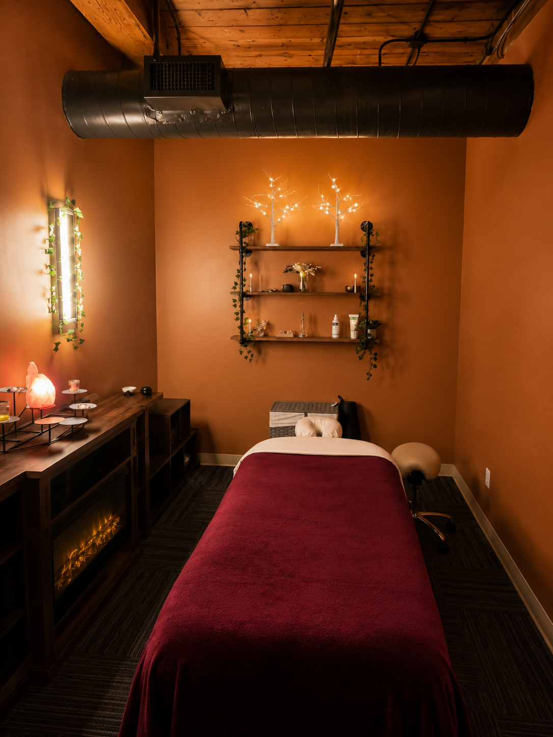 A massage room with a bed covered in a burgundy blanket, warm orange walls, ambient LED lighting, decorative shelves with plants and candles, and a large black industrial vent on the ceiling.