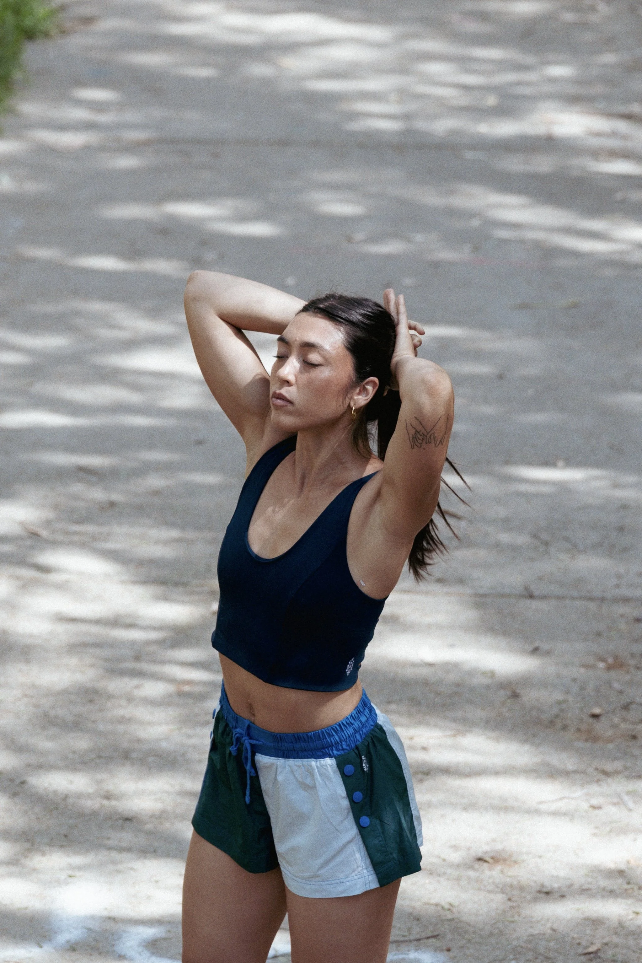 A woman in workout clothes standing outdoors with closed eyes, hands behind her head, and expression of relaxation.