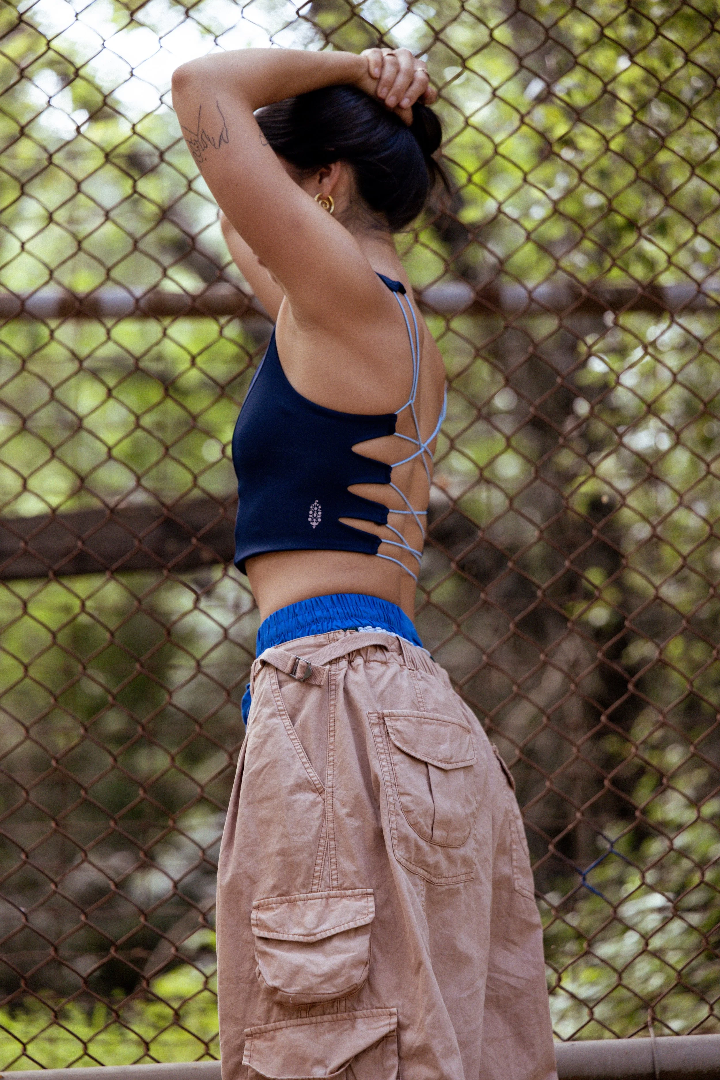 A person standing in front of a chain-link fence with greenery, wearing a blue tank top and beige cargo pants, with their arms raised and hands behind their head.