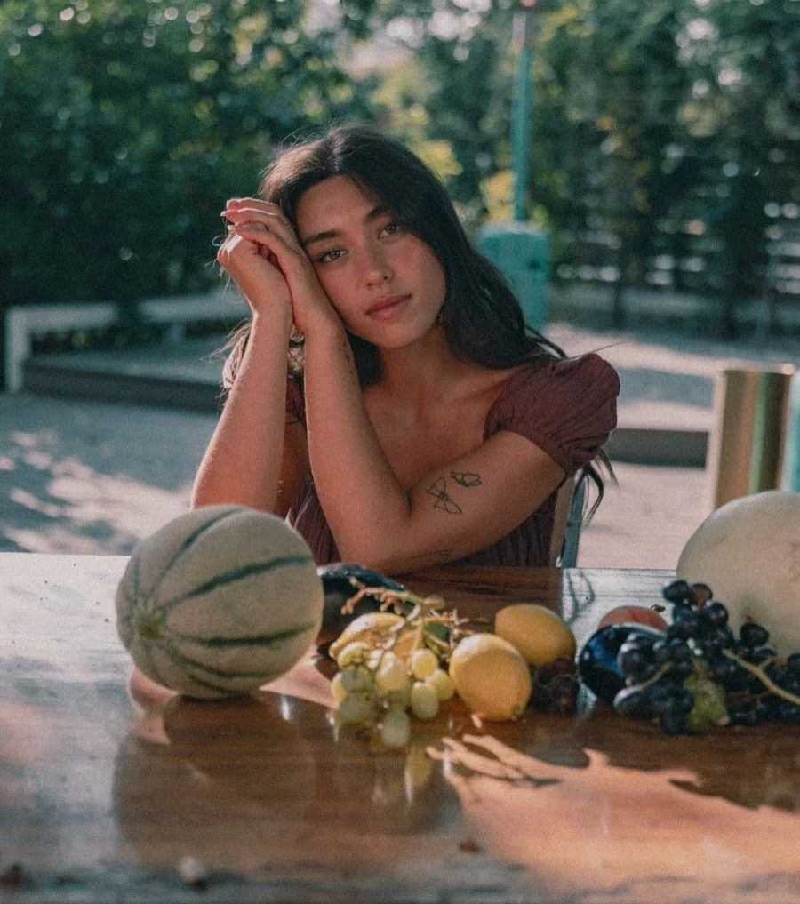 A young woman with long dark hair sitting at a wooden table with various fruits, including a melon, grapes, and lemons, outdoors on a sunny day, with green trees in the background.