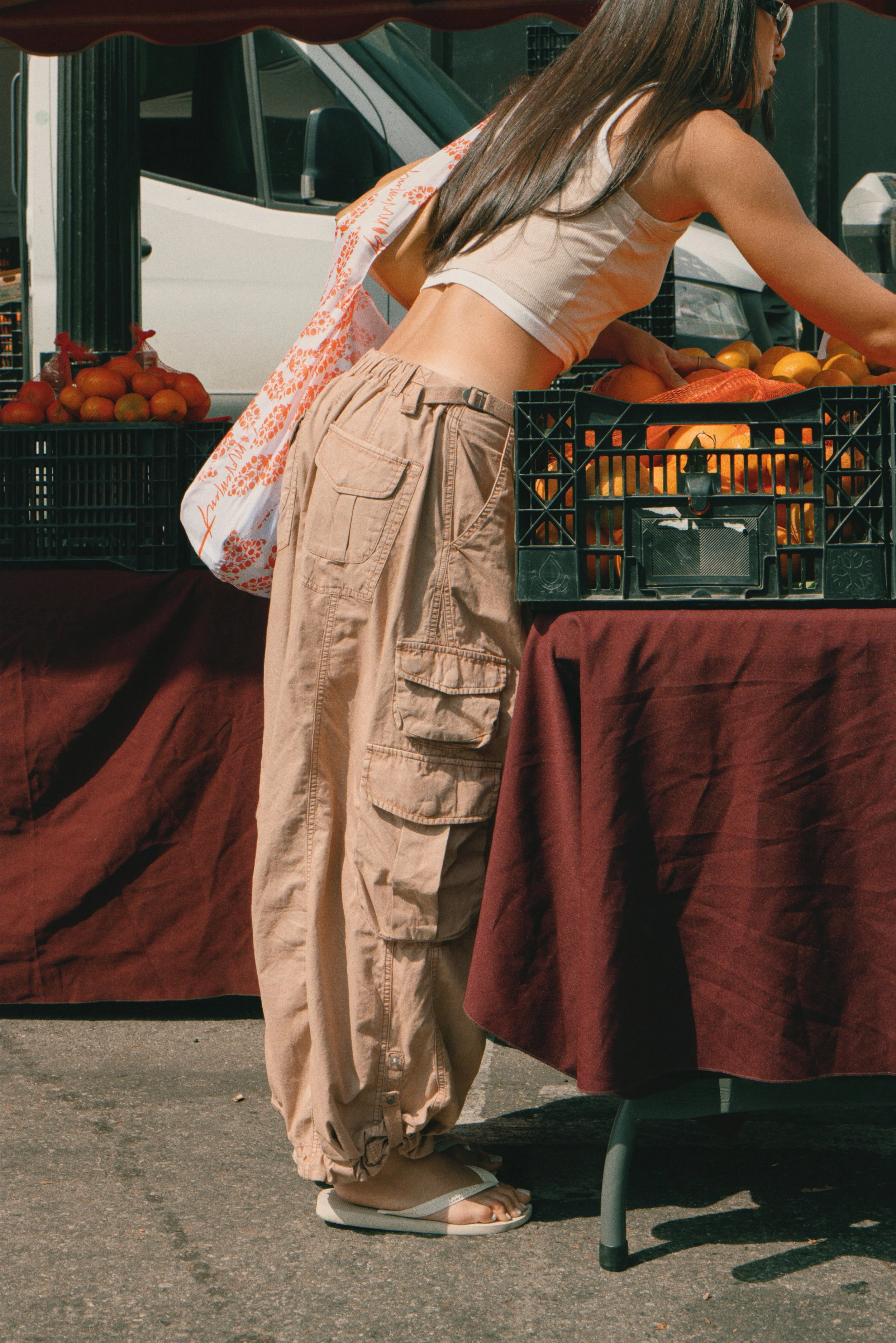 A woman at a farmer's market wears tan cargo pants, a beige crop top, and white sandals, selecting produce from a table with oranges and other fruits.