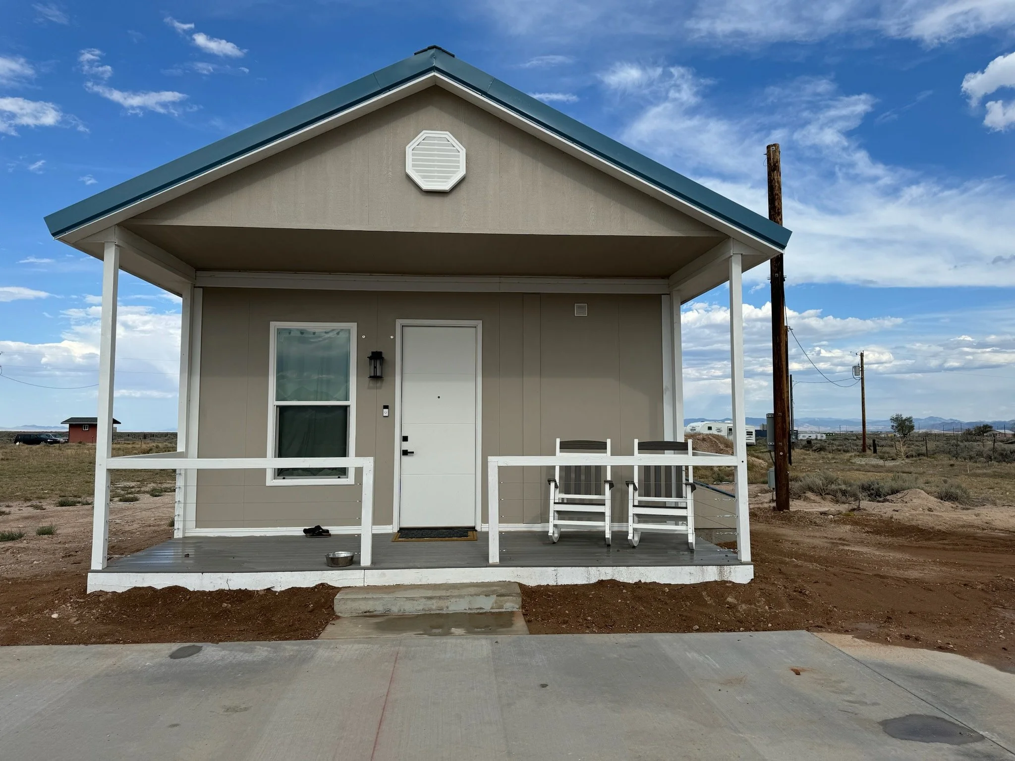 Small beige house with a front porch, white railing, and two chairs on the porch. The house has a sloped blue roof, one window, and a white door. The house is in a barren landscape with a dirt ground and a partly cloudy sky.