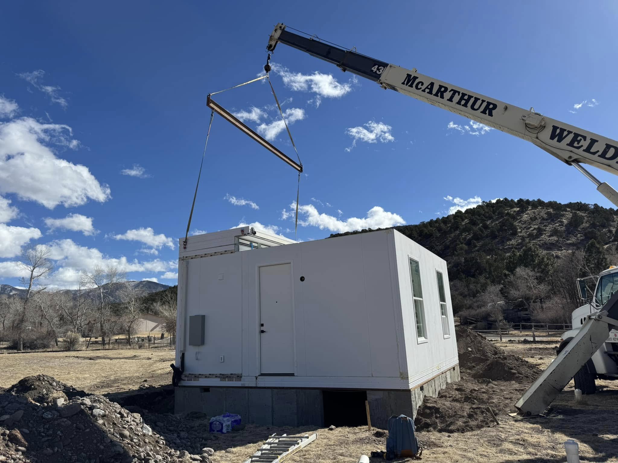 A white modular building being lifted by a crane with a blue sky and mountains in the background.