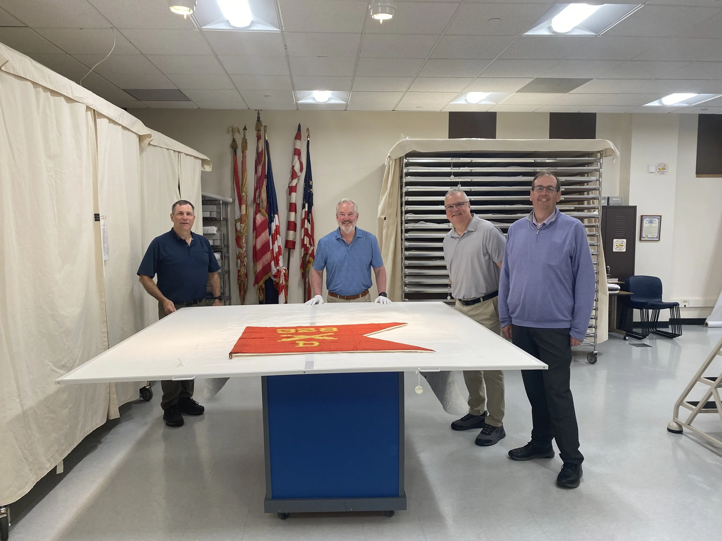 Four men stand behind a small triangular shaped, red flag laying on a table. The flag reads 328 D and has crossed cannons. To the left and right are tall cases covered in canvas.