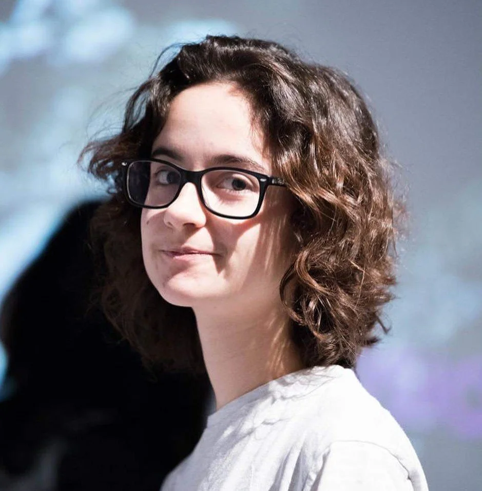 A young woman with curly brown hair, glasses, and a white shirt stands outdoors, smiling slightly and looking towards the camera with a blurred background.