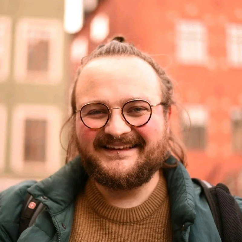 A young man with glasses, a beard, and a mustache, smiling outdoors in front of a brick building.